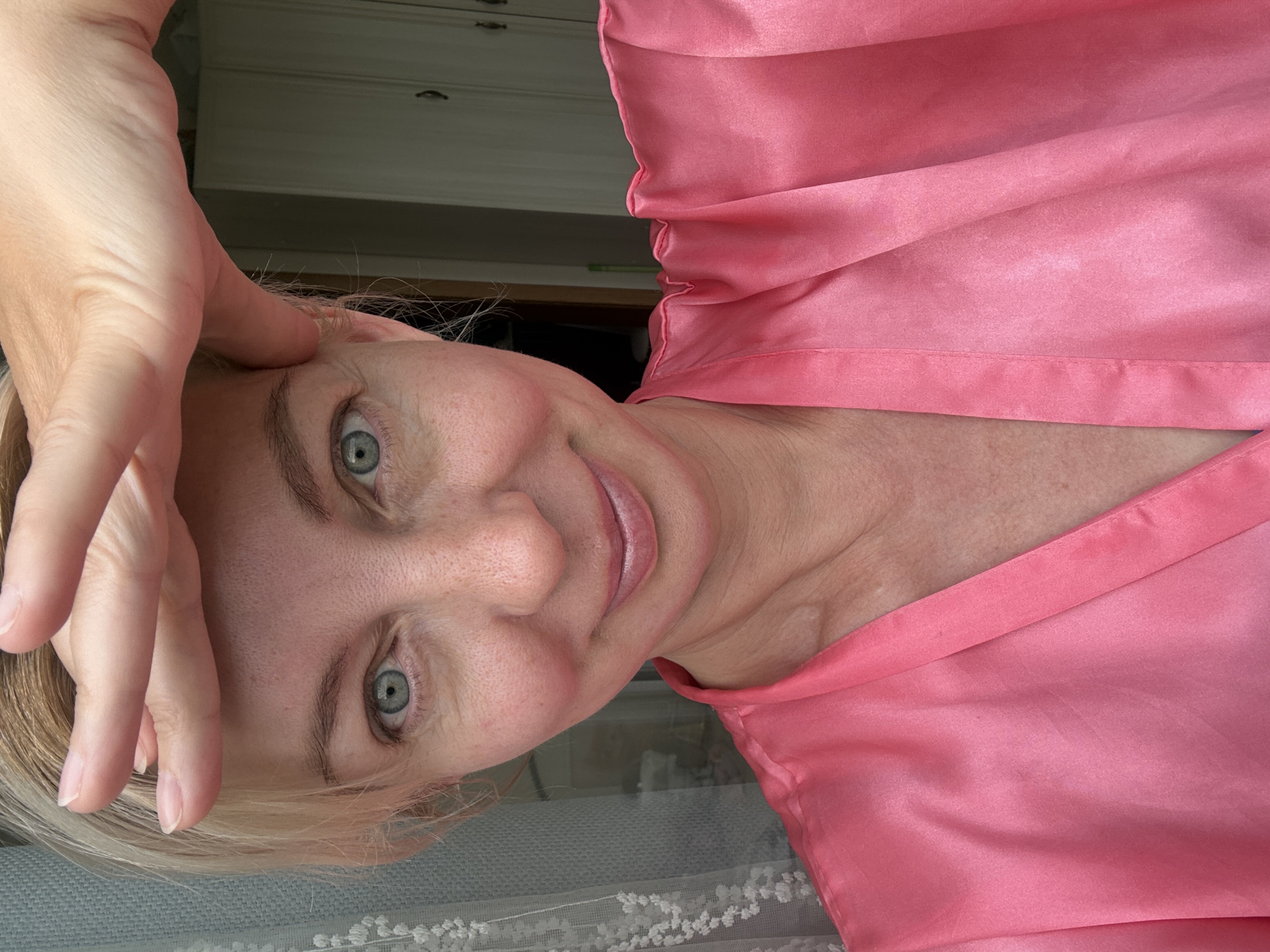 A woman with blonde hair and blue eyes, wearing a pink robe, poses for a selfie. Her right hand is raised to her forehead, shielding her eyes from the sun. She is smiling slightly, and her lips are painted with a light pink lipstick. The background shows a window with a lace curtain on the left and a closet with two doors on the right. The setting appears to be an indoor space, possibly a bedroom or dressing room. There is no information in the image to suggest it is located in Sochi, Russia.