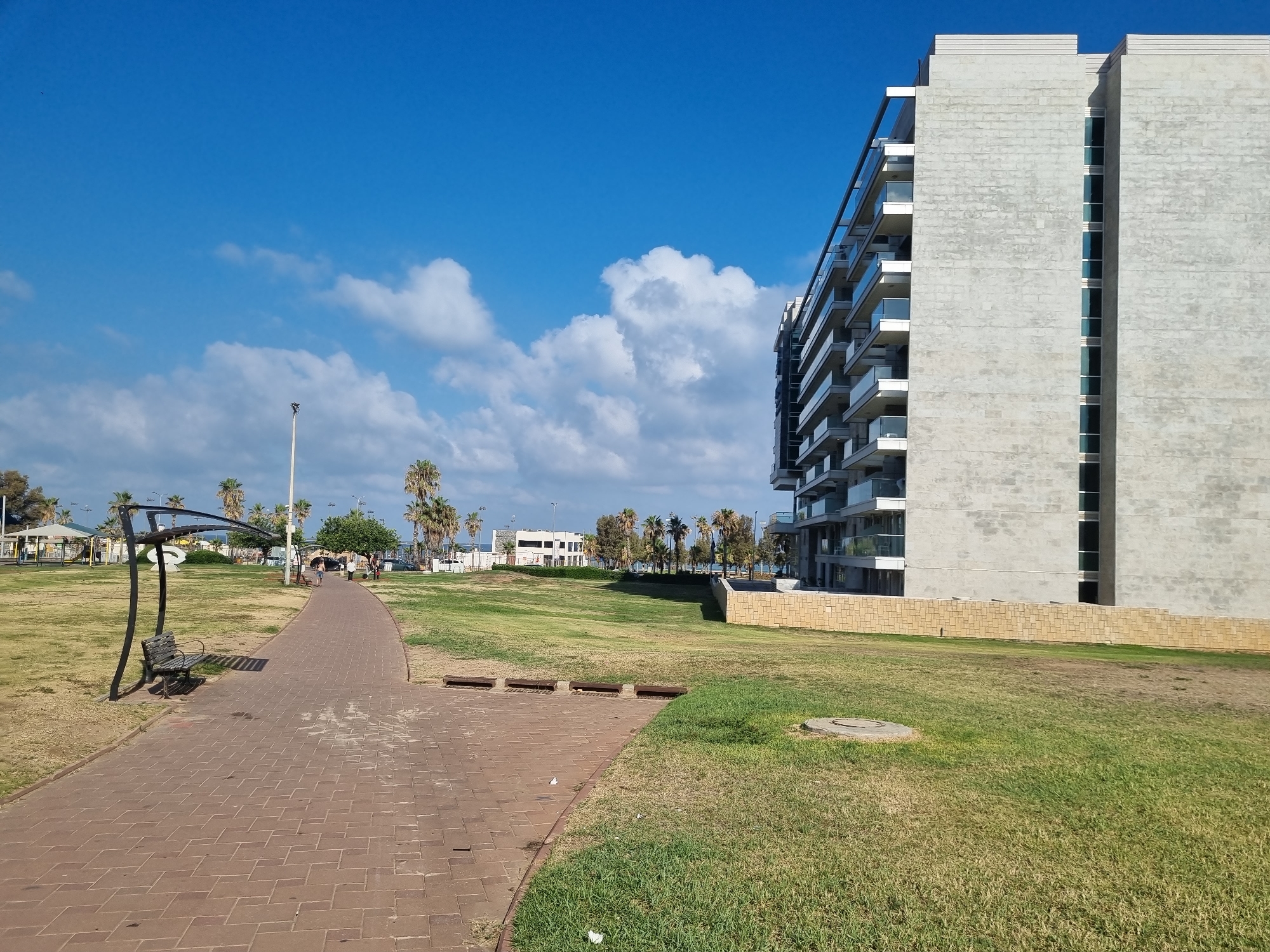 The image depicts an outdoor scene in Ashdod, Israel, featuring a modern building, a park area with a paved walkway, and a bright blue sky with scattered white clouds. The building, likely an apartment complex, is tall and has a contemporary design with multiple balconies and glass railings. It is situated on the right side of the frame, partially obscuring a low, light-colored stone wall.
In the foreground and extending to the left, a red brick walkway curves through a grassy park. A solitary bench with a dark metal frame and wooden slats sits beside the path, casting a shadow. Further down the walkway, a few individuals are visible, walking away from the camera. The park is mostly covered in green grass, with some patches appearing dry. Palm trees and other trees are scattered throughout the park, with some appearing to be in the distance. In the far left of the image, a playground structure with a white roof and an open-sided metal frame can be seen. The overall impression is one of a well-maintained public space on a clear day.