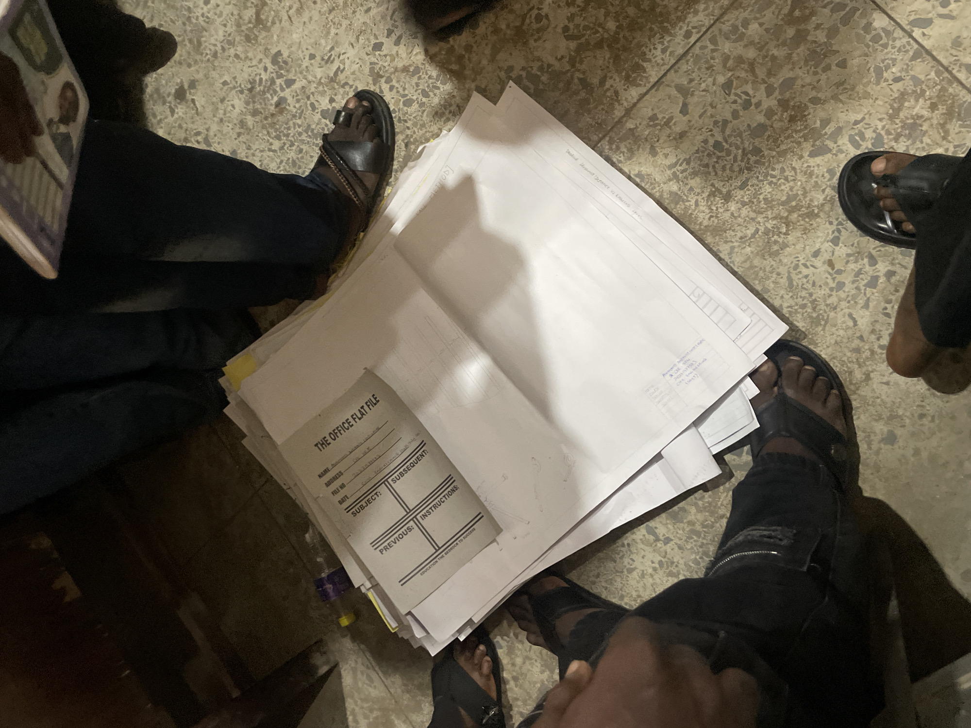 In Ihiagwa, Nigeria, a pile of documents is spread across a speckled floor. The documents include a gray file folder labeled "THE OFFICE FLAT FILE," with fields for "NAME," "ADDRESS," "FILE NO," "DATE," and "SUBJECT." Some lines are filled in with handwritten text. Beneath the file folder are several white sheets of paper, some with handwritten notes and diagrams. Four individuals are partially visible in the frame, their legs and feet clad in dark trousers and sandals. One person is holding a book or magazine. The environment appears to be an indoor setting, possibly an office or a home, with a dark wooden structure visible in the background. The lighting is dim, creating shadows on the floor and highlighting the textured surface.