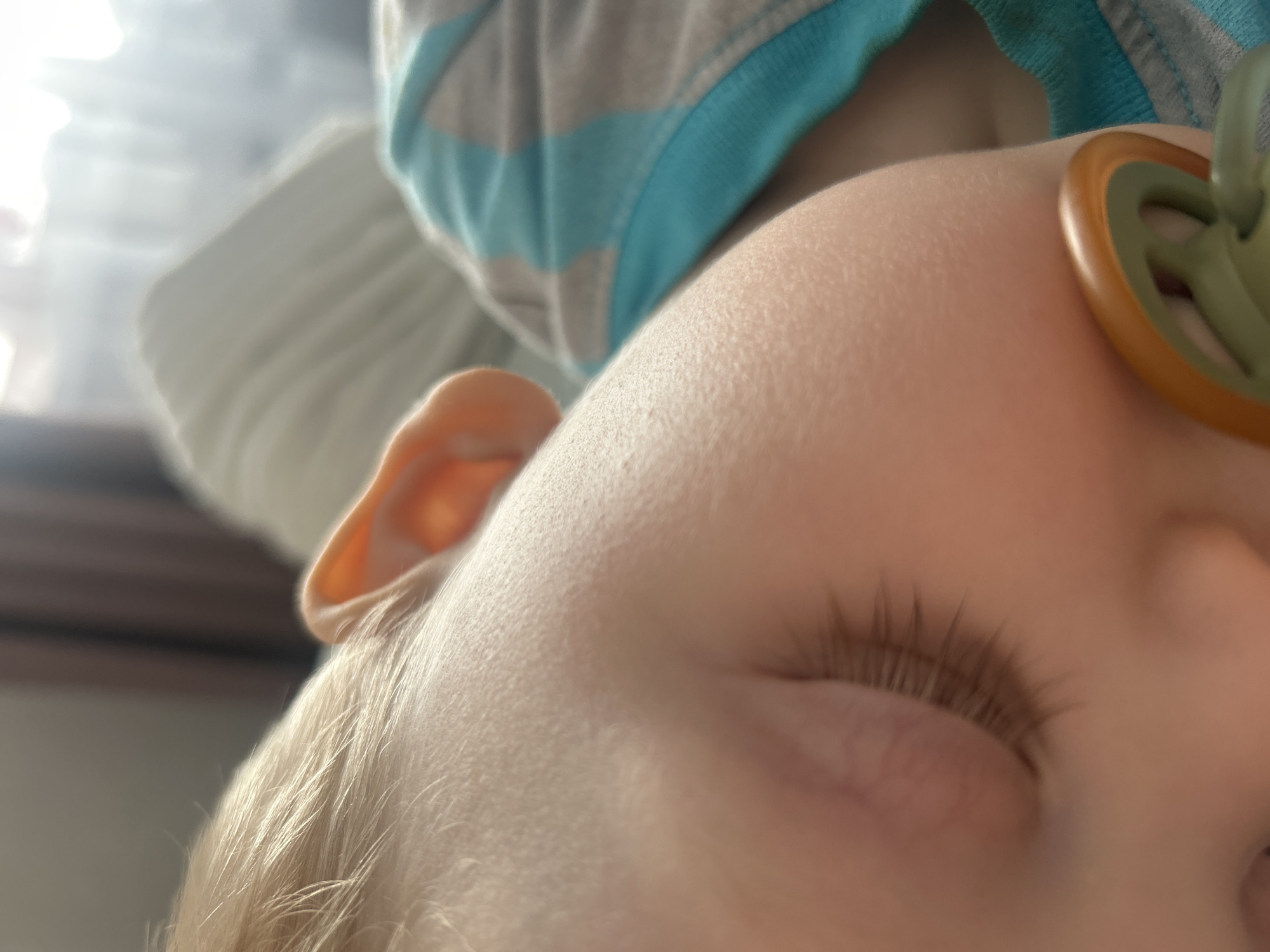 A close-up, slightly blurred image depicts the upper right side of a sleeping infant or toddler's head, positioned with the face generally pointed towards the left of the frame. The child exhibits fair skin, fine light-colored hair, and closed eyes with distinct eyelashes. A pacifier, featuring a green nipple and an orange-brown circular outer rim, is partially visible near the child's mouth and cheek area. The child is dressed in a garment with alternating light blue and light grey or white horizontal stripes. The background is out of focus, showing a diffused, bright light source in the upper right quadrant, consistent with a window, and blurred light-colored bedding or fabric. A darker, indistinct shape is present in the upper left background. The scene is located in the city of Chaikovsky, Russia.