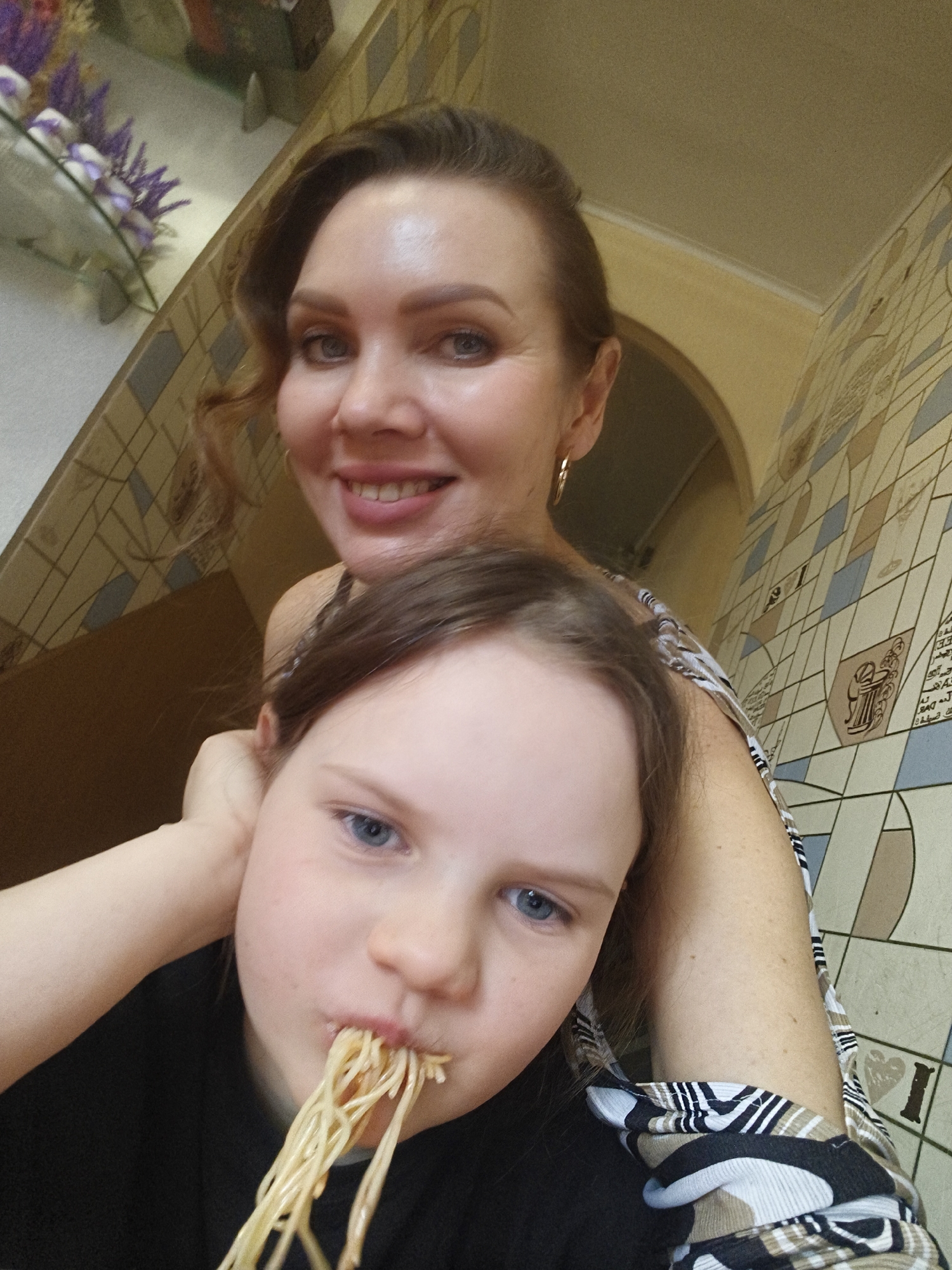 The image shows a selfie of a woman and a child indoors in Unknown, Unknown. The woman is smiling and has brown hair pulled back. She is wearing earrings and a patterned top. The child is in the foreground, with spaghetti hanging out of their mouth. They have light hair and blue eyes, and are wearing a dark top. The background includes patterned wallpaper and a glimpse of a shelf with purple flowers.