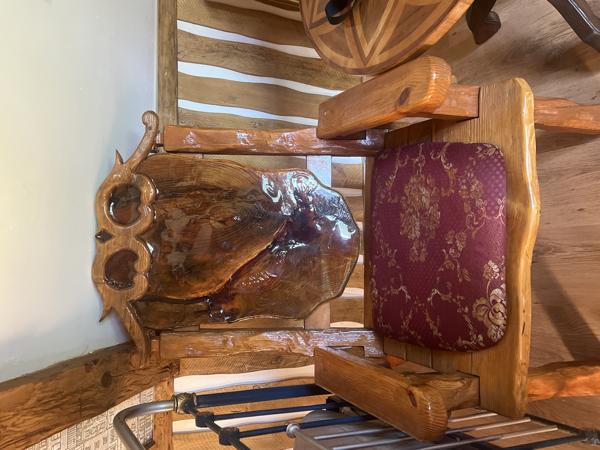 The image shows a handcrafted wooden chair with a carved owl design on the backrest. The chair has a cushioned seat with a red fabric featuring a gold floral pattern. The chair is placed indoors, with wooden paneling on the wall behind it. A portion of a circular wooden table with an inlaid geometric pattern is visible to the right of the chair. The lighting suggests an indoor setting, possibly a home or a rustic establishment. The style of the chair and the surrounding decor imply a natural or artisanal theme. The image does not contain any information about the location in Daulėnai, Lithuania.