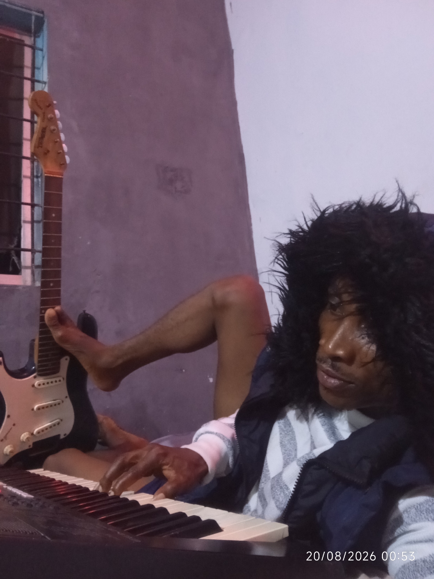 The image shows a person in Adankolo, Nigeria, positioned with their legs extended while playing a keyboard. To their left, an electric guitar is propped upright. The person is wearing a black hooded jacket with fur trim over a striped sweater. The environment appears to be an indoor space with a muted purple wall in the background and a window with metal bars. The lower right corner of the image displays a timestamp: 20/08/2026 00:53.