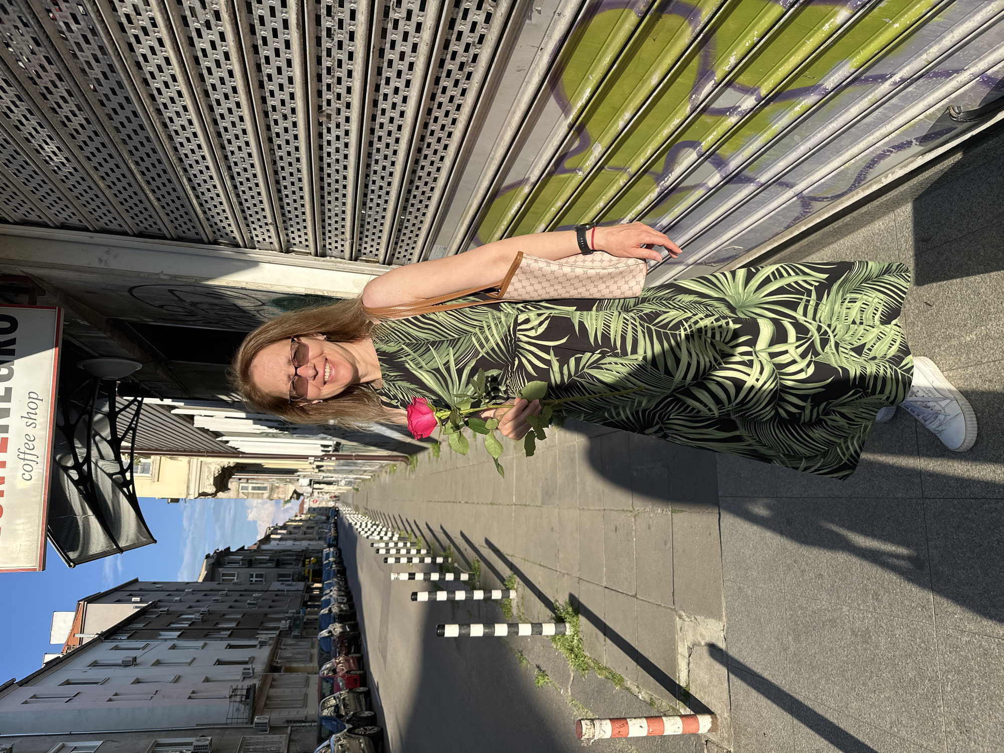 A woman stands on a sidewalk holding a pink rose. She is wearing a black and green palm-leaf print dress, white sneakers, and sunglasses. She has a tan and pink patterned tote bag on her shoulder. Behind her is a closed storefront with a green metal roll-up door covered in graffiti. To her left is a street lined with parked cars and bollards. In the background, buildings with balconies and air conditioning units line the street. The scene is located in Sofia, Bulgaria, as indicated by the Cyrillic text on the coffee shop sign.
