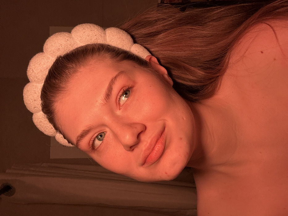 A fair-skinned woman with blonde hair is tilted slightly to the right. She has a white headband with a bubbly texture. Her eyes are light and she gazes to the left. The light in the image gives off an orange tint. The background has a vague shape of a door and bed.