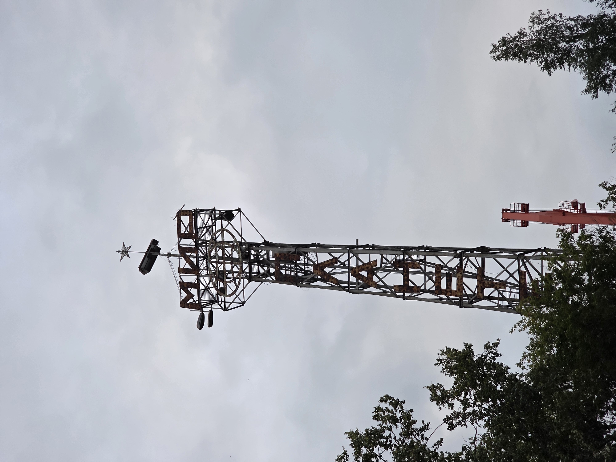 The image depicts a tall, rusty metal tower with the word "МИР" (Mir, meaning "peace") prominently displayed at its top in large, weathered Cyrillic letters. Below this, more Cyrillic letters, possibly forming another word or phrase, are visible and appear to be vertically aligned along the tower's structure. The tower is topped with a star. In the background, a dull, overcast sky dominates the frame. To the left of the main tower, the lush green foliage of trees partially obscures the lower section. In the lower right quadrant, another, smaller red structure, possibly a part of another tower or a different piece of industrial equipment, is visible, along with more trees. The overall impression is one of an aging, possibly abandoned, Soviet-era monument or structure against a somber sky. The location is identified as the city of Chaikovsky, Russia, suggesting this is a landmark within the city. The image captures a static, outdoor scene with no visible people or animals.