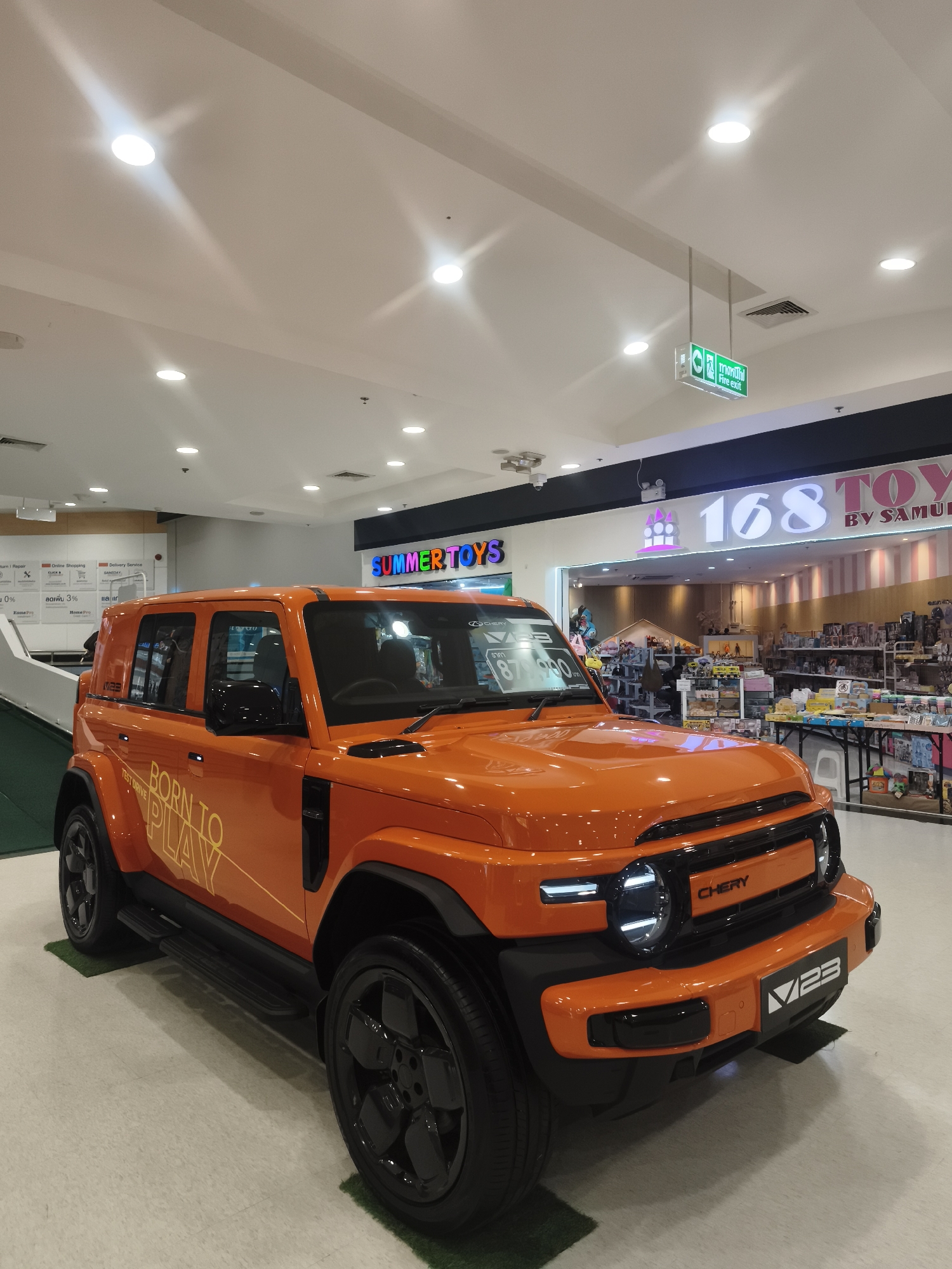 An orange Chery V23 SUV is displayed in an indoor setting, possibly a shopping mall, in Ko Samui District, Thailand. The vehicle is positioned on a light-colored tiled floor, with two small artificial grass mats placed under the front and rear tires. The car features black wheels, side steps, and fender flares. "BORN TO PLAY TEST DRIVE" is visible on the side of the vehicle. Behind the car, a store with a bright pink and white striped facade and a sign reading "168 TOYS BY SAMUEL" is visible, suggesting a retail environment. The ceiling features recessed lighting and a green "Fire Exit" sign. No people are visible in the image.