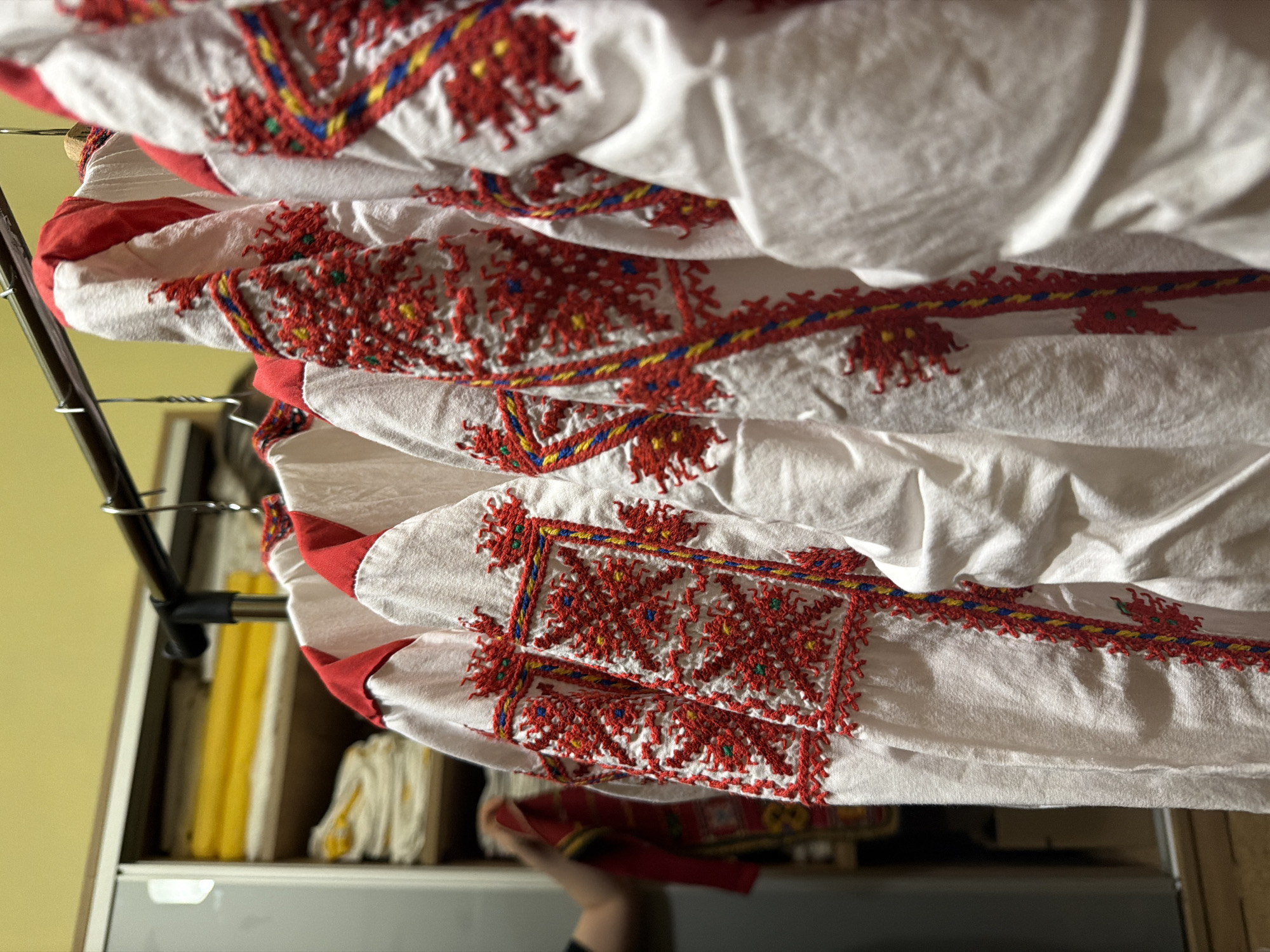 The image depicts several white cotton shirts with red embroidered designs, hanging on a metal clothes rack. The shirts appear to be traditional Bulgarian folk costumes, possibly from the region of Sofia, Bulgaria, based on the style of embroidery. In the background, a shelf unit contains various items, including yellow and white textiles. A person's hand is partially visible at the bottom of the image, holding a red piece of fabric. The overall lighting suggests an indoor setting, possibly a home or a storage area.
