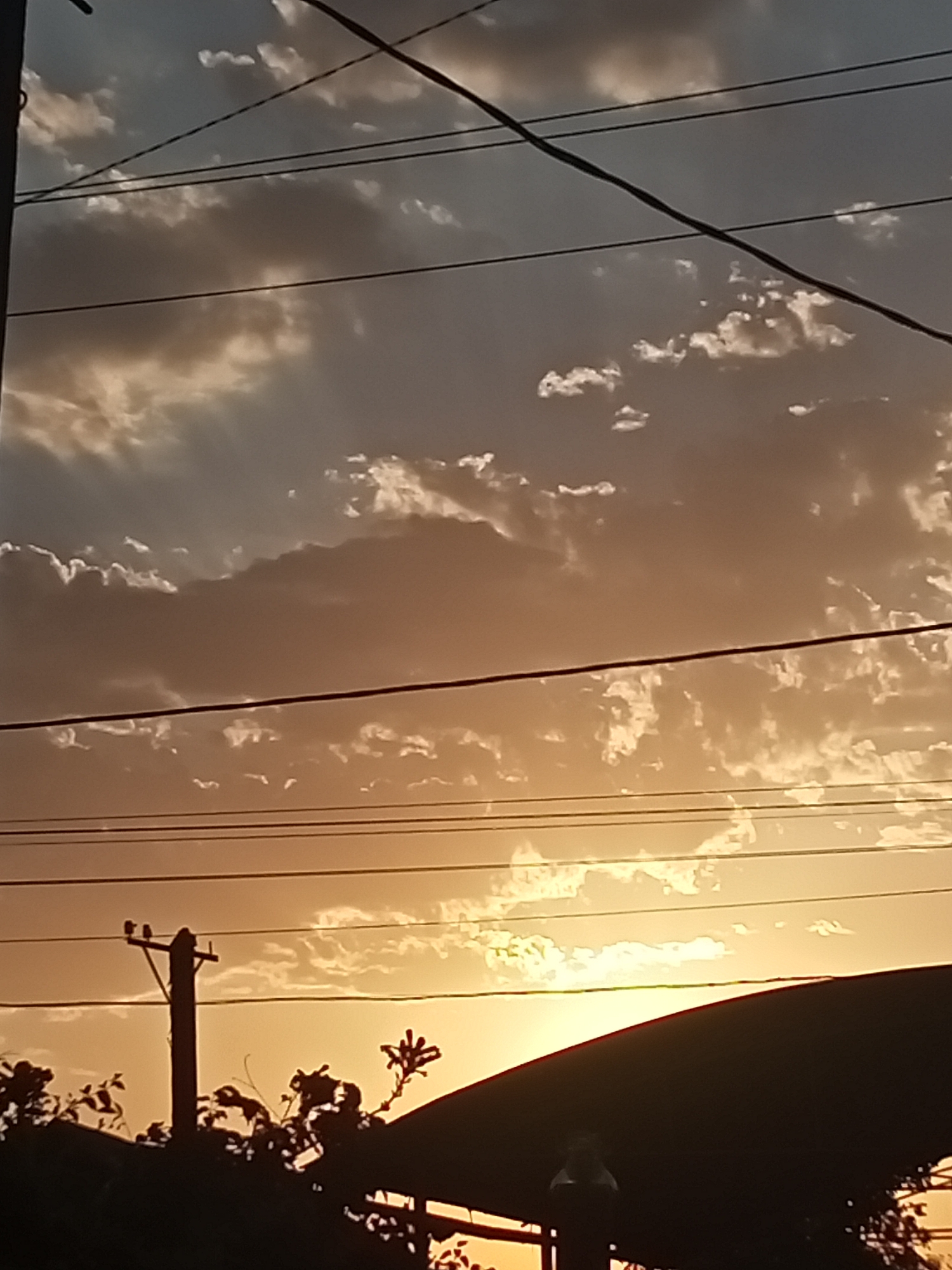 A silhouette of a utility pole with wires and some dark foliage is visible against a dusky sky with scattered clouds. The sun is setting, casting a warm orange glow across the sky. The image appears to be taken outdoors, possibly in a semi-urban or rural area of Sokuluk, Kyrgyzstan.