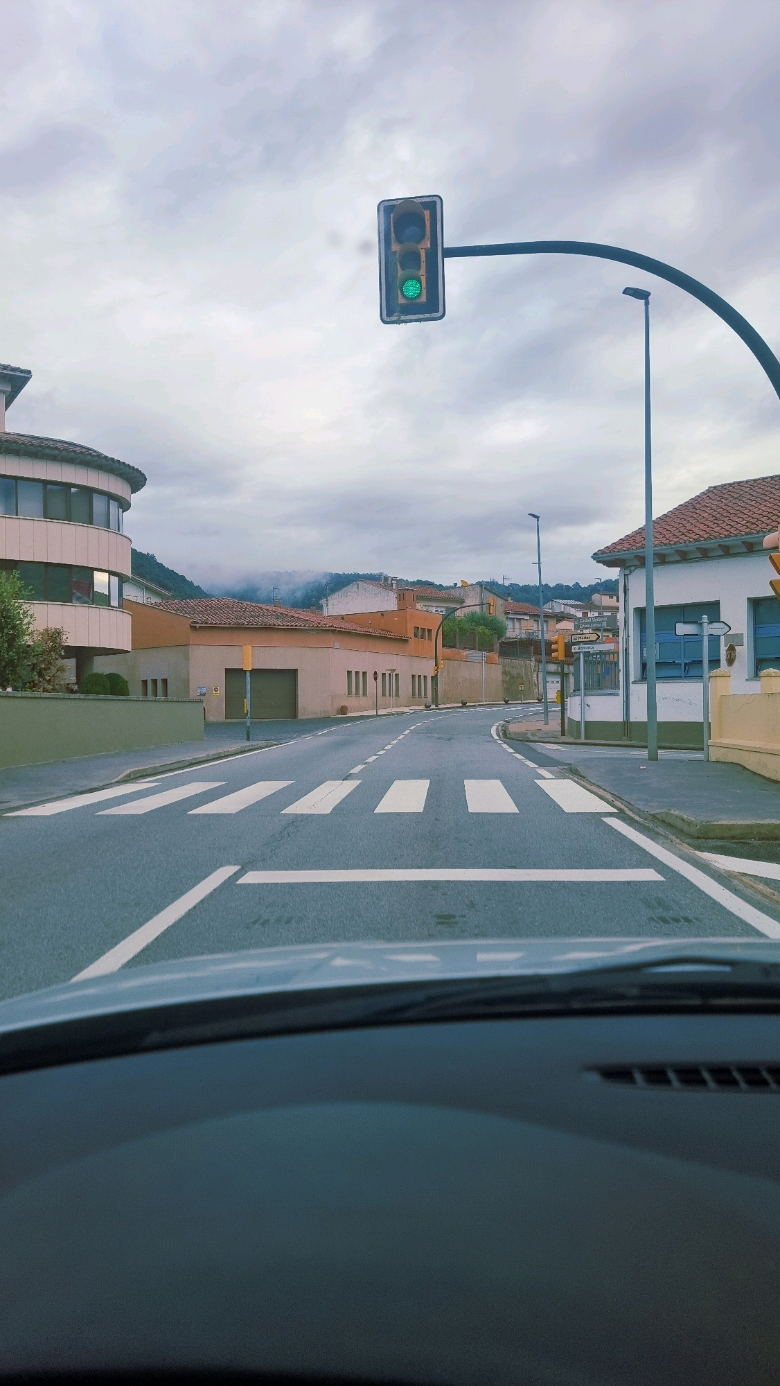 The image shows a view from a vehicle looking towards a pedestrian crossing on a street in Sant Joan Les Fonts, Spain. A traffic light is illuminated green, indicating that traffic can proceed. The street is paved with asphalt and marked with white lines. Buildings line both sides of the street, with one building on the left having a curved facade and several windows. On the right, a building with a red tile roof is visible. The sky is overcast with clouds. In the background, hills can be seen. The scene appears to be captured during daylight hours.