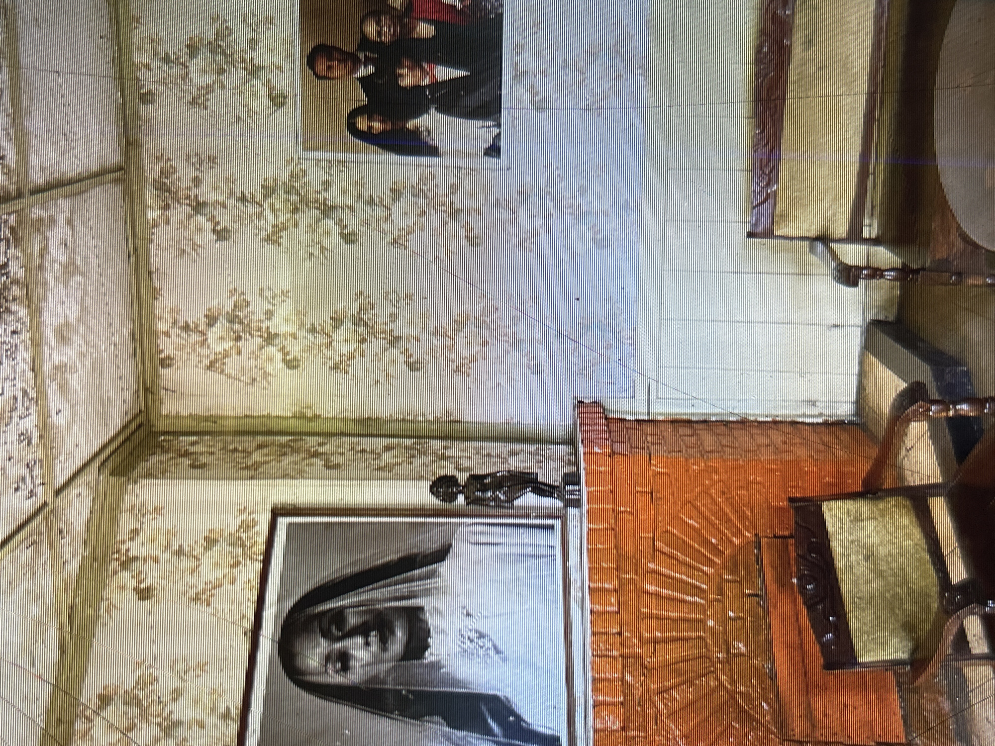 The image depicts an interior scene, likely a living room, characterized by vintage decor. On the left, a framed portrait of a woman in a veil hangs above a brick fireplace. To the right of the portrait, a small, dark statue stands on the mantelpiece. Further to the right, another framed photograph displays a family of four. In the foreground, a wooden armchair with yellow upholstery sits to the left, while a wooden bench with similar upholstery is positioned to the right. The walls are adorned with floral wallpaper, and the ceiling also appears to have a textured pattern. The lighting suggests an indoor setting, possibly during the day. There are no overt indications of the city of Depok, Indonesia within the visual elements of the image itself.