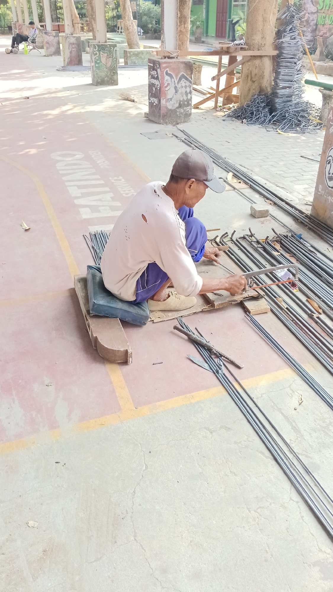 In Branggahan, Indonesia, an elderly man with a worn-out shirt and blue pants sits on a blue cushion on a wooden plank on a pink asphalt surface. He is using a hacksaw to cut through metal rebar. Several bundles of metal rebar lie on the ground around him, some bent into various shapes. In the background, there are concrete pillars decorated with paintings, trees, and wooden structures. A person is visible sitting on a chair in the far background. The scene appears to be outdoors, possibly on a sports court or a similar open area.