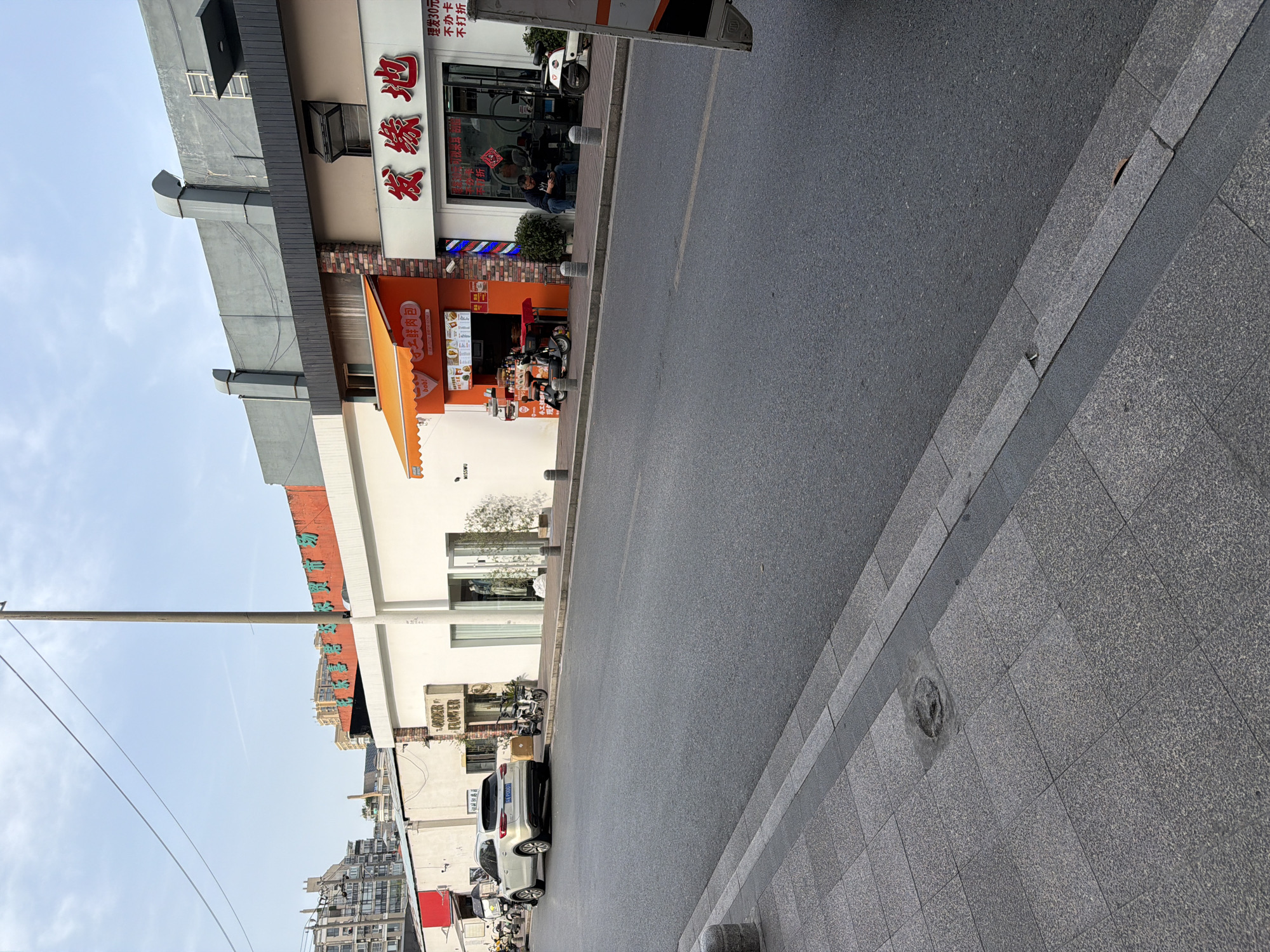 The image captures a street scene in Jiangning District, China. The perspective is from the street level looking toward buildings lining the road. The street itself, made of asphalt, occupies a significant portion of the frame. A sidewalk consisting of textured tiles runs alongside the street. Several businesses are visible along the buildings, including what appears to be a hair salon named "发缘地" with Chinese lettering and a barber pole. A small eatery labeled "工鲜肉包" with the word "bob!" is also identifiable by its orange awning and signs. Cars and scooters are parked along the curb. A power line stretches across the upper left portion of the frame against a light blue sky.