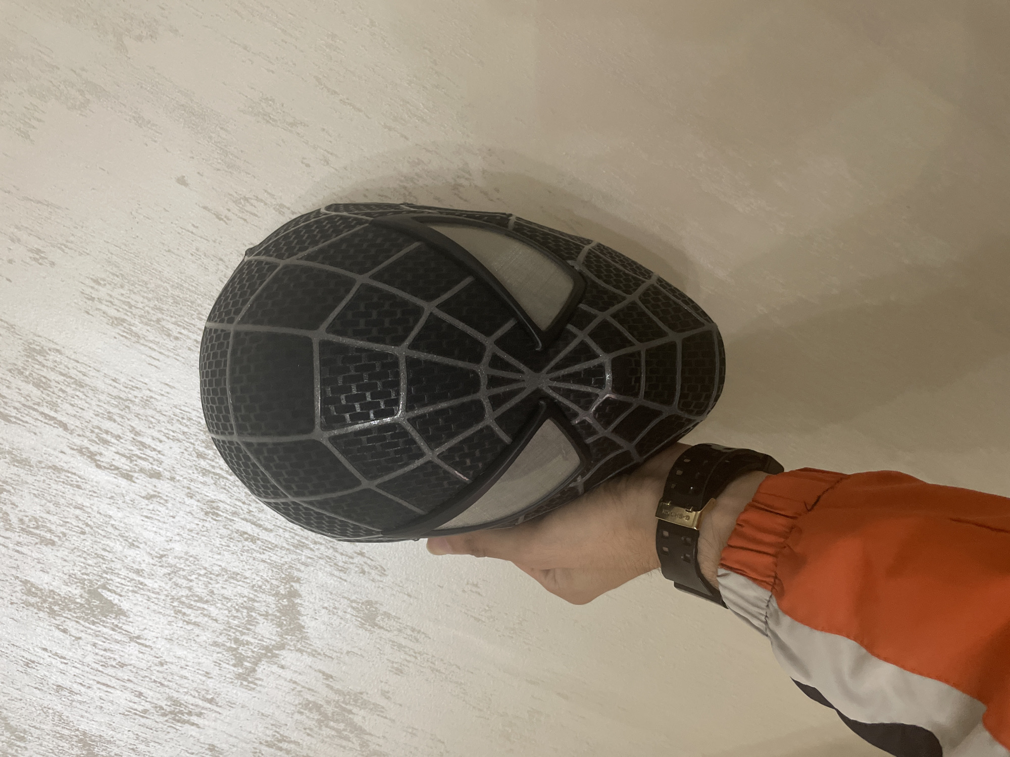 The image, captured from an elevated perspective, shows a human hand with a light skin tone holding a black Spider-Man mask against a textured, off-white wall in the city of Ihtiman, Bulgaria. The mask, styled after the Symbiote suit, features a black base with silver-grey web-like lines and large, almond-shaped eye sections with silver-grey borders. The mask exhibits a rigid structure, with the black areas displaying a fine, woven texture. The hand is positioned towards the lower-left, gripping the mask from beneath its left side. A black wristwatch with a gold buckle is secured around the wrist. The sleeve of a multi-colored garment, featuring prominent orange with white and black segments, extends from the forearm.