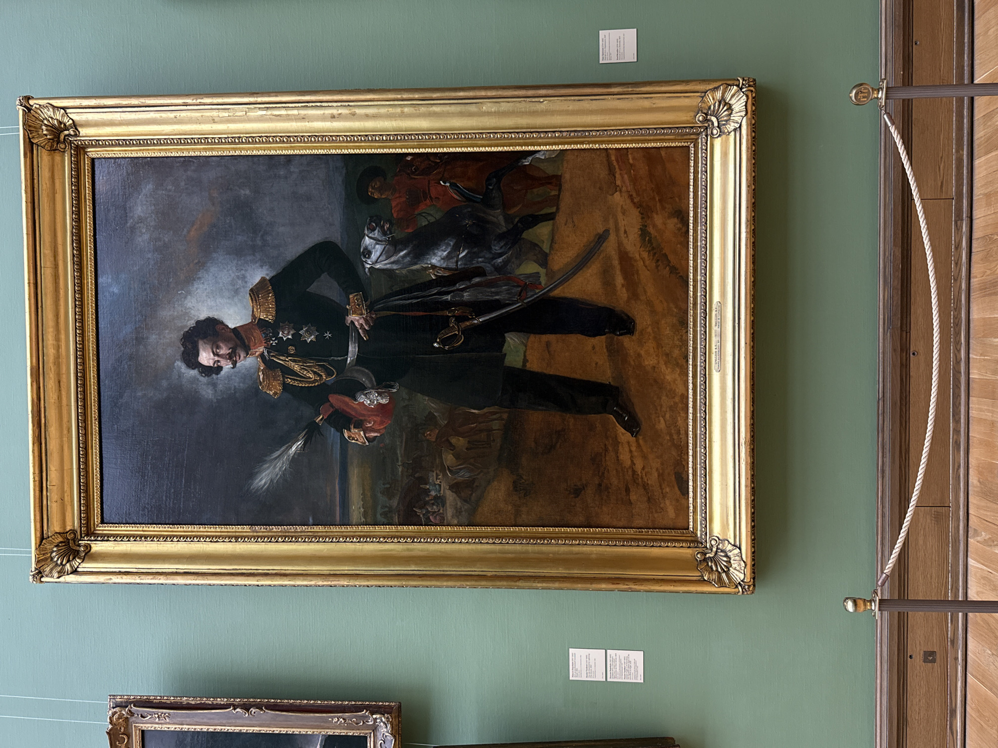 A framed portrait painting hangs on a green wall. The painting depicts a man in a military uniform with gold epaulettes and multiple medals, standing next to a horse. There's a man riding the horse, and other figures in the background. The frame is ornate and gold-colored. A rope and a metal bar are visible next to the painting, suggesting it's in a museum. The image was taken at the State Tretyakov Gallery in Moscow, Russia.