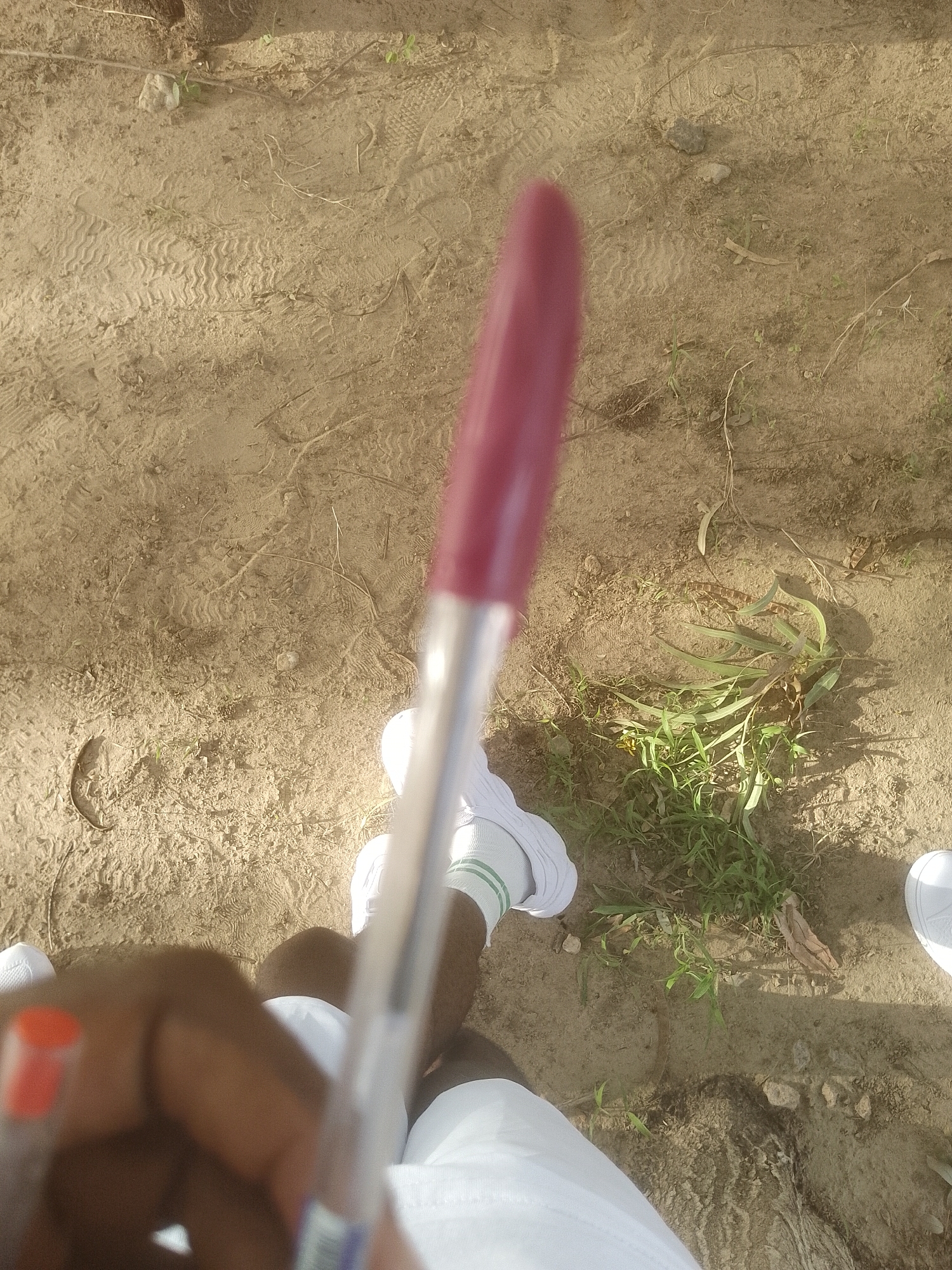 The image shows a first-person perspective of someone holding a red-capped pen with their left hand, which is partially visible. The pen is held at an angle, obscuring the background in the foreground. The individual is wearing white socks with green stripes and white sneakers. Other visible elements include sand, dirt, scattered dry grass, and some green plants. There are also shoe prints visible in the sandy ground. In the background, more of the sandy terrain and some greenery are present. The location is identified as Majemu, Nigeria.