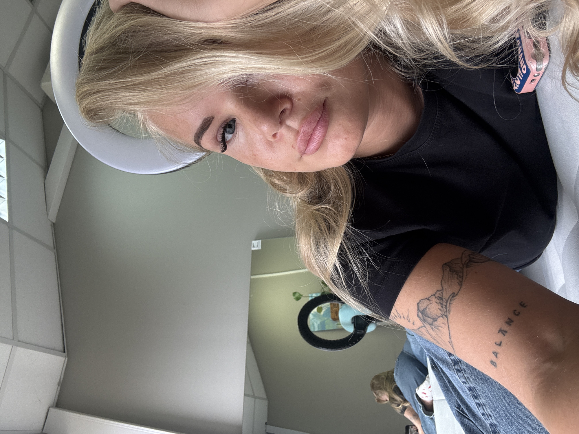 A close-up, slightly off-kilter photo features a young woman with blonde hair wearing a black t-shirt. She appears to be under a ring light, indicated by the white circular shape above her head. The woman has fair skin, light eyebrows, and pink lips. Her left arm is visible and displays two tattoos: one depicting mountains and another spelling "BALANCE" vertically.
In the background, there's a glimpse of a room interior. The walls are light gray. A mirror hangs on the wall, showing a reflection of a plant and another person, possibly seated. The location is in the city of Unknown, Unknown.