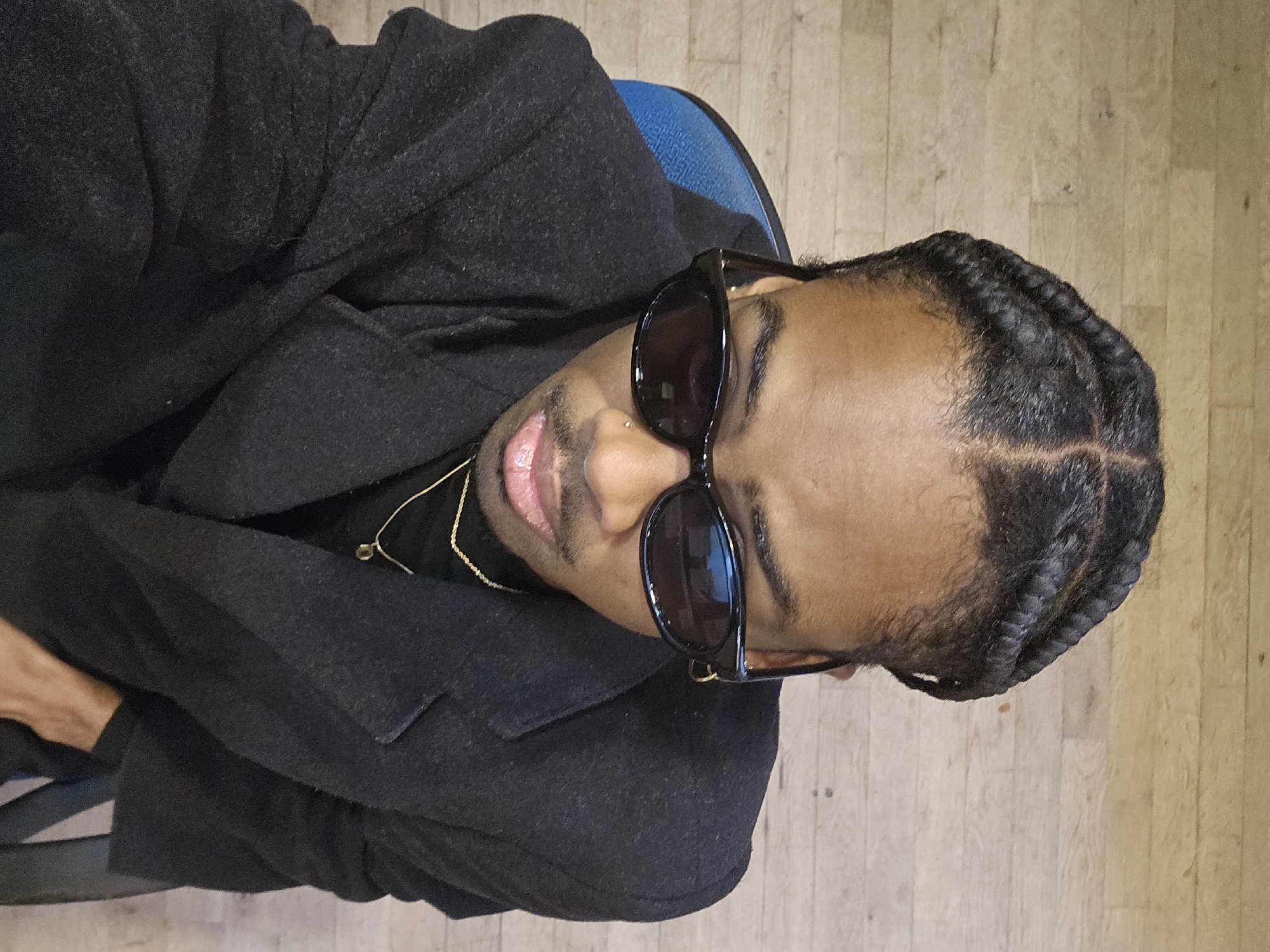 A man with braided hair is wearing sunglasses and a dark coat while sitting on a blue chair. The location appears to be indoors, with a light wooden floor. There is no indication in the image that the location is in Paris, France.