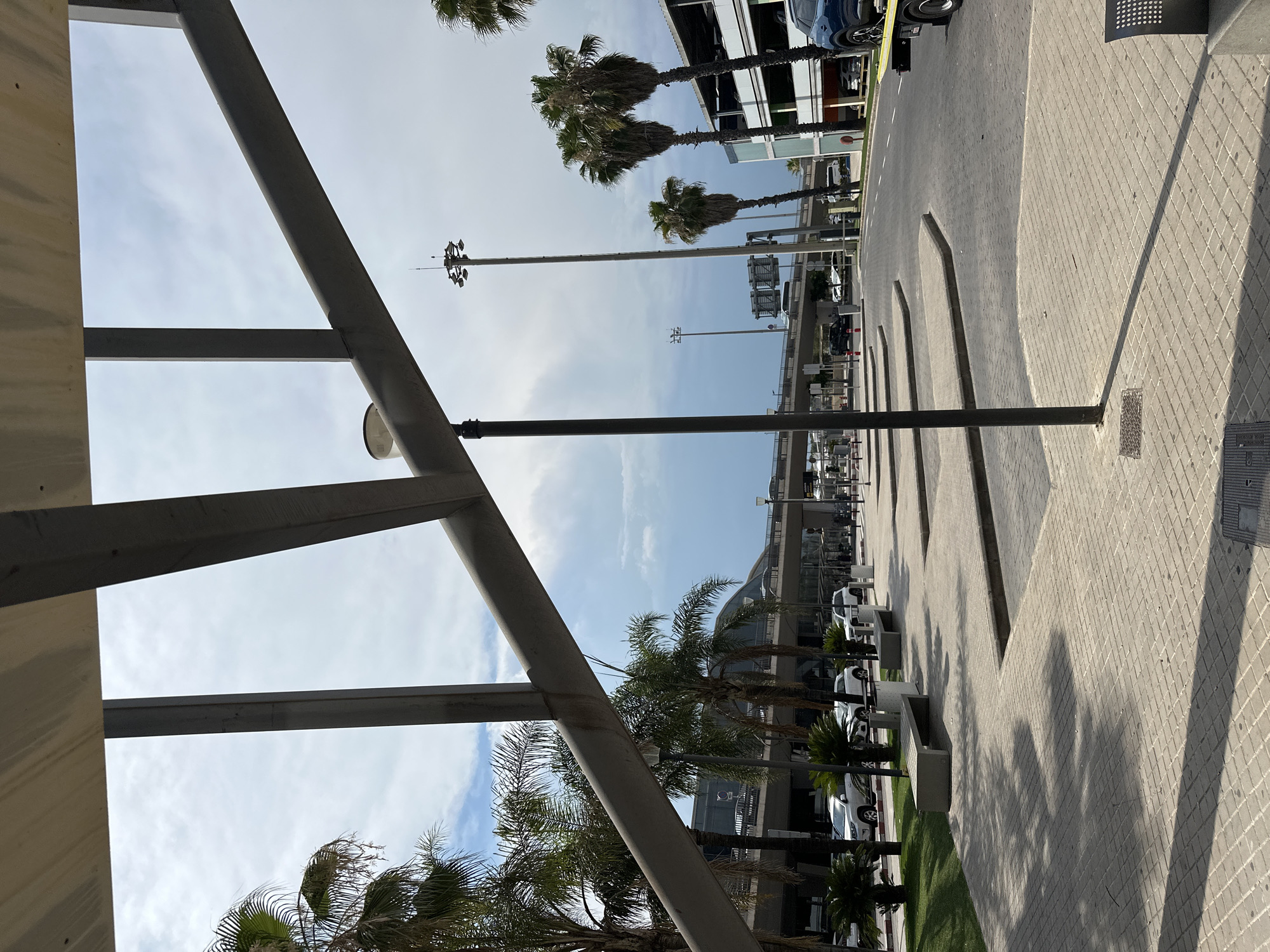 The image depicts an outdoor scene with a focus on architectural elements and a parking area. In the foreground, a large metal structure with diagonal and horizontal beams dominates the left side of the frame, suggesting a modern canopy or walkway. The structure casts shadows on the ground.
Beyond the metal structure, a paved area is visible, laid with grey square tiles arranged in a diamond pattern. This area appears to be a pedestrian walkway or part of a plaza. Several raised concrete planters are lined up along the edge of the paved area, some with greenery or small palm trees. Further down the walkway, there are multiple cars parked, indicating it's a parking lot or a street. The cars are of various colors, including white and dark blue.
In the background, a modern building with large windows and a curved facade is visible, possibly a commercial building, airport terminal, or public facility. Palm trees are scattered throughout the scene, adding a tropical or Mediterranean feel. A tall lamppost with multiple lights stands in the middle ground. The sky is partly cloudy, with patches of blue sky and white clouds, suggesting daytime. The lighting indicates it is likely mid-morning or afternoon. The overall impression is a well-maintained public space in a warm climate, possibly near a transportation hub or a resort area. No people are clearly visible in the frame. No text is discernible in the image.