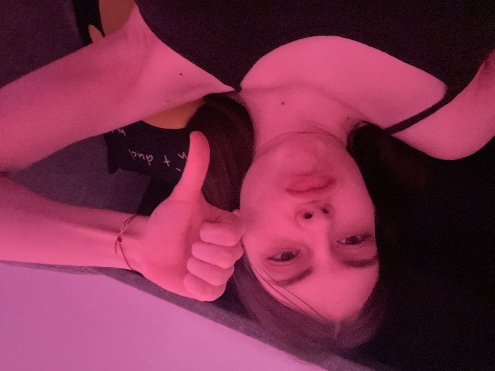 A person with fair skin, dark hair, and a neutral expression is lying down and giving a thumbs up. They are wearing a black top with spaghetti straps. A thin red bracelet is visible on their wrist. A patterned black and yellow object is in the background. The lighting is predominantly pink. The location is not discernible and therefore not identifiable as Saint Petersburg, Russia.