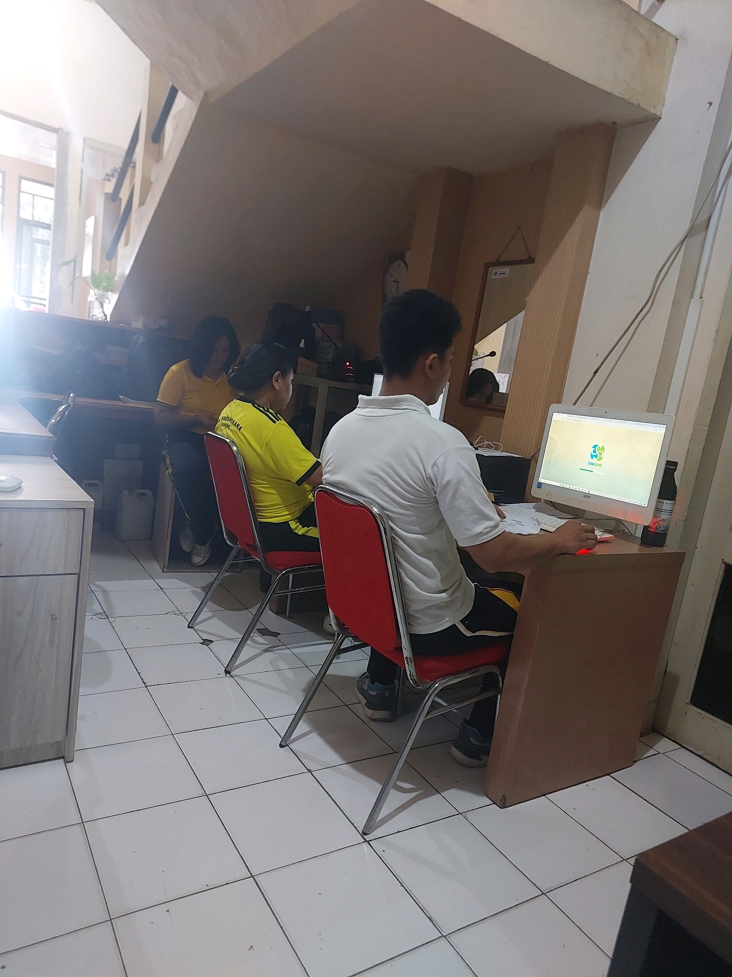 The image shows an indoor scene of what appears to be an office or computer lab, possibly located in Kupang, Indonesia, given the style of the building and interior. In the foreground, a man is seated at a desk, facing a computer monitor displaying a green interface with a logo. He is wearing a white polo shirt and dark shorts. To his left, a red chair is visible. In the background, two women are seated at desks, also facing computers. Both women are wearing yellow shirts. One woman in the middle ground is facing away from the camera and appears to be looking at her computer. The other woman, to her left, is also looking at her computer. The environment includes tiled flooring, a staircase in the background, and various office furniture and equipment. The lighting suggests it is daytime, with natural light coming from a window on the left side of the frame, creating some glare. The overall impression is a functional workspace.