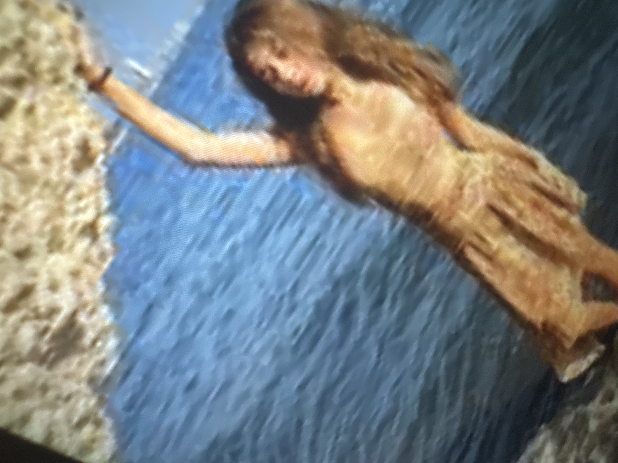 This blurry and low-resolution image captures a person, likely a young woman, with long, flowing brown hair, floating or swimming in a body of water. She is positioned diagonally across the frame, with her head facing slightly towards the viewer and her left arm outstretched towards a sandy or rocky bank on the left side. The woman appears to be wearing a light, possibly sandy-colored, dress or garment that blends with her skin tone.
The setting is an outdoor body of water, possibly a river, lake, or ocean, adjacent to a landmass. The water is blue with visible ripples or gentle waves, suggesting movement. The overall activity depicts the person calmly floating or swimming near the shore. The perspective is from above, looking down at the individual in the water. The lighting suggests it is daytime, with bright conditions. No specific emotions are clearly discernible due to the image quality and distance. There is no visible text in the image.