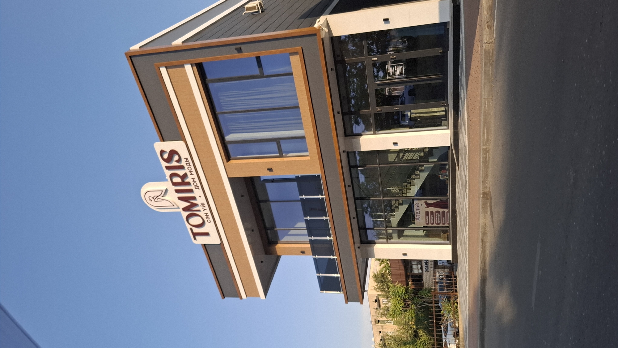 The image shows a two-story building with the sign "TOMIRIS" prominently displayed on the upper level. Below the main sign, smaller text reads "сән үйі" and "дом моды," which translates to "house of style" in Kazakh and "fashion house" in Russian, respectively. The building has large windows on both floors, a balcony with glass railings on the second floor, and an entrance on the ground floor. A sign inside the ground floor entrance reads "Ателье" (Atelier), indicating a tailoring or dressmaking service. Several cars are visible parked on the street in front of the building and a sidewalk with decorative paving stones. The building is located in Shymkent, Kazakhstan, as indicated by the text on the signage. The sky is clear and blue, suggesting it is daytime.