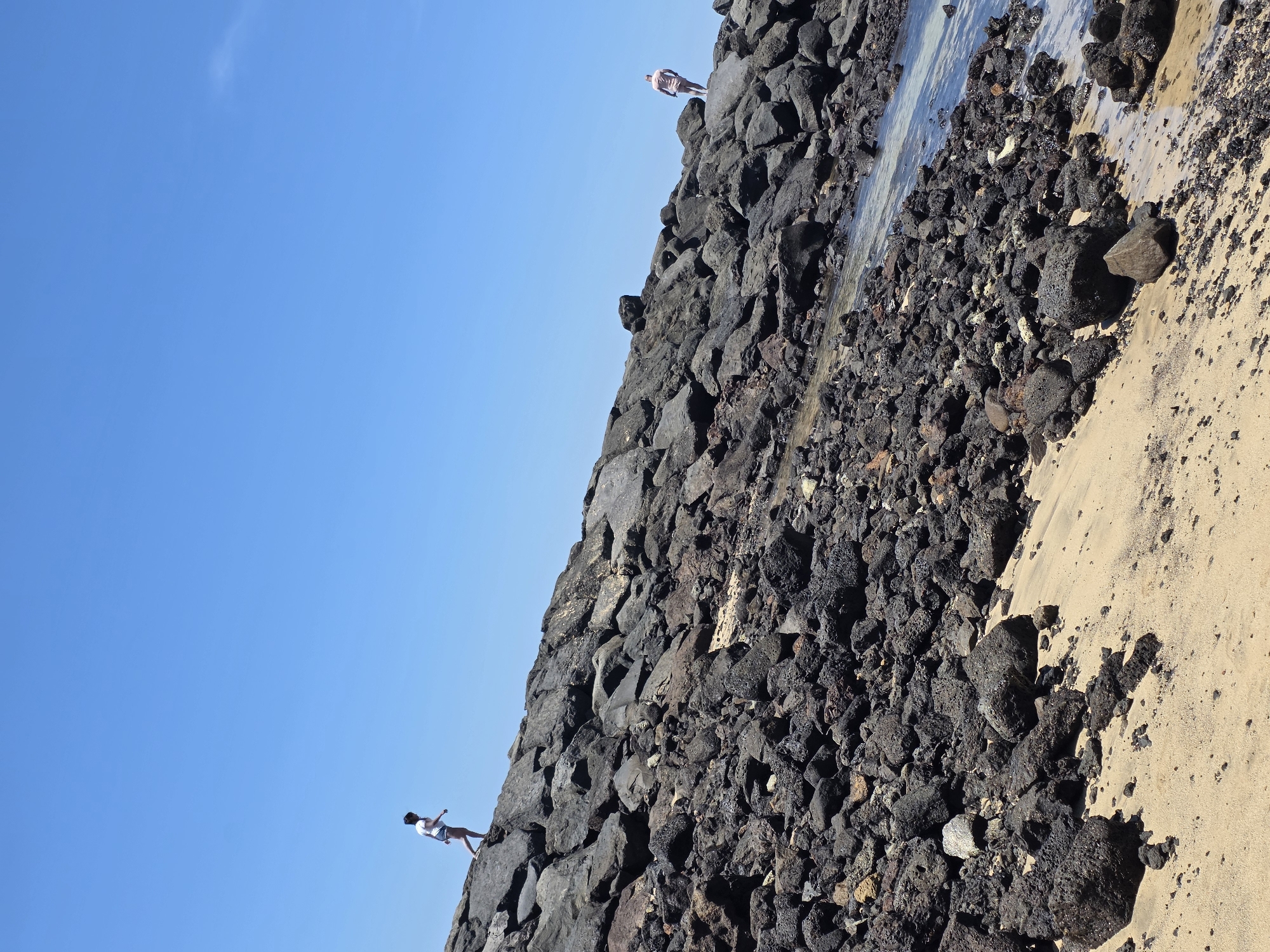 The image shows a sunny day at the beach in Costa Teguise, Spain. Two individuals are visible on a rocky coastline. On the left, a person wearing a white t-shirt and shorts is walking along the top of a large rock formation. Further to the right, another person in light-colored shorts is standing on a rock overlooking the water. The foreground consists of sand and numerous dark, irregular rocks, with a small area of shallow water on the right edge of the frame. The sky is a clear, bright blue.
