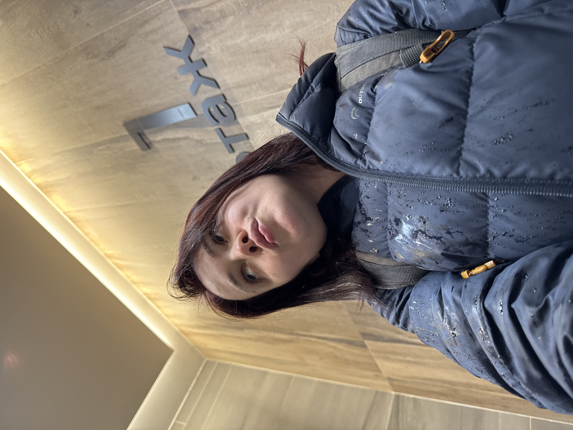 An upward-looking portrait captures a person, likely female, with dark, possibly reddish-brown hair, wearing a dark blue puffer jacket. The jacket appears wet with numerous small water droplets or has a reflective, speckled texture. A backpack strap is visible over their right shoulder. Faint text reading "OUTD" and a partial crescent-shaped logo are visible on the jacket, suggesting outdoor apparel. The person's lips are slightly pursed, and their expression is neutral.
The setting is indoors, likely a modern commercial or institutional building in Izhevsk, Russia. The background features light-toned, possibly tiled or paneled walls and ceiling. A bright, linear architectural light strip runs along a corner. Prominently displayed on the wall/ceiling behind the person is a metallic sign with bold letters. The visible text on this sign clearly reads "TeXK". The image is taken from a low angle, giving an unconventional, rotated perspective where the person and the background elements appear tilted. The wet appearance of the jacket could indicate the person has recently entered from outside.