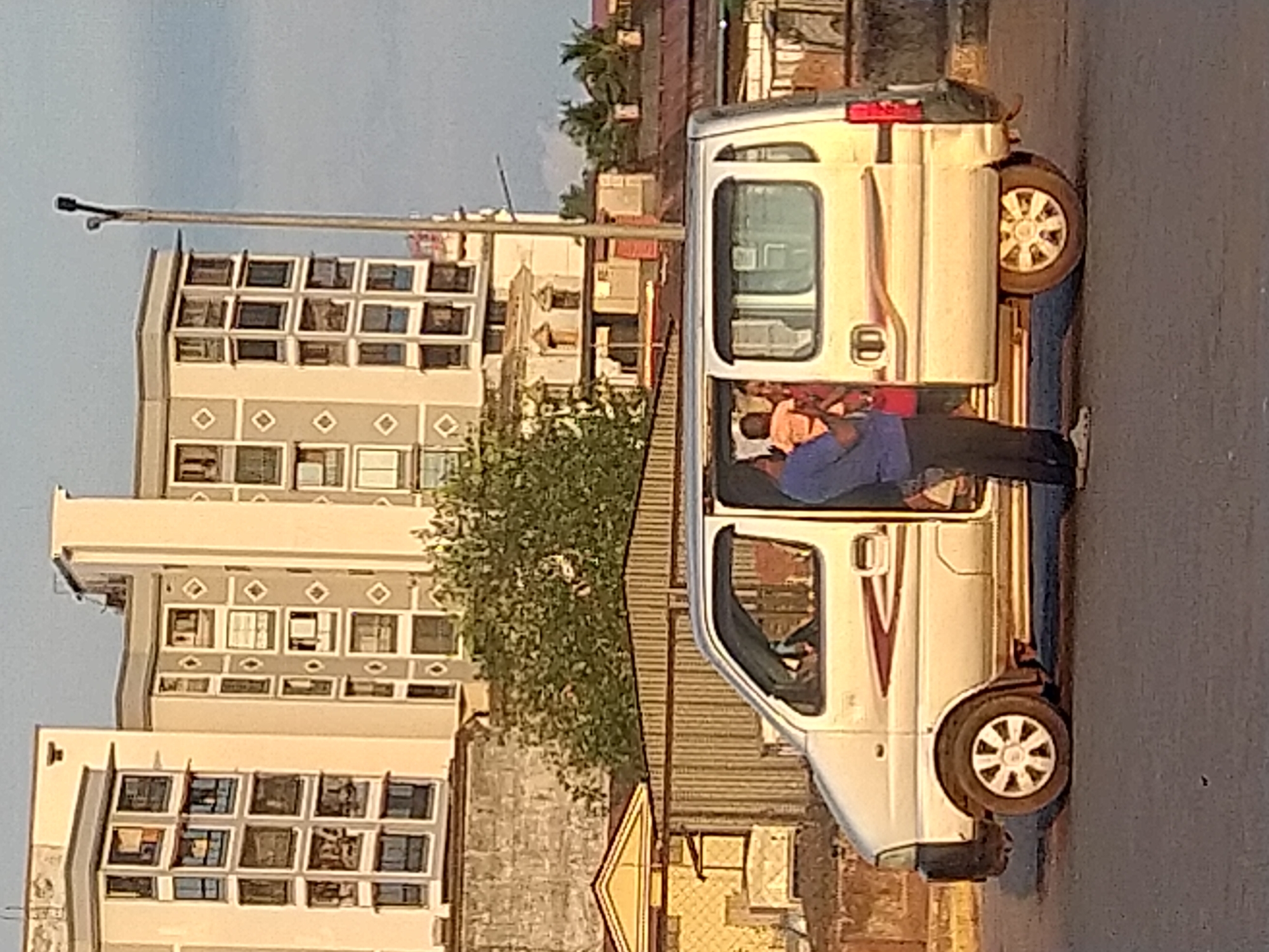 The image shows a silver van parked on a street in Mapusa, India. A man in a blue shirt is standing partially inside the open driver's side door, seemingly speaking to passengers within. The background includes a building with multiple windows, a street light, and some foliage. The setting appears to be an urban area.