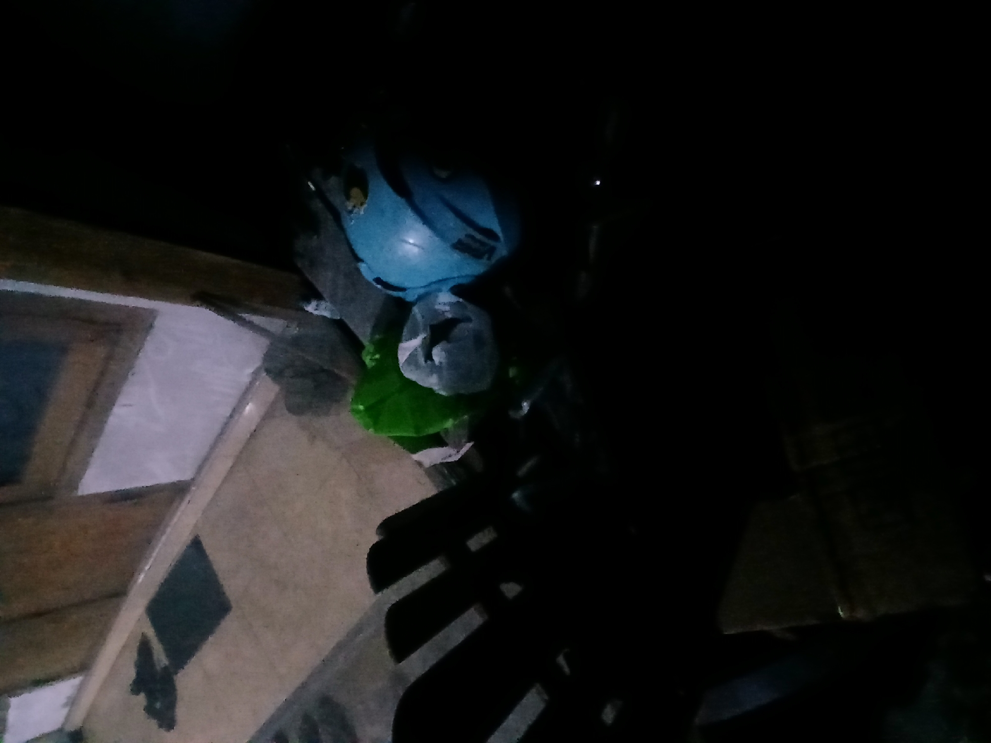 The image depicts an outdoor scene at night, likely in the city of Kademangan Kulon, Indonesia. In the foreground, there is a dark, slatted object, possibly a chair, casting shadows. To the right, a blue helmet rests on a surface along with some green and white material, possibly plastic bags. In the background, a doorway or entrance is visible with a wooden door and a white tiled wall. Several dark-colored shoes are placed on a doormat in front of the entrance. The scene is dimly lit, creating a dark and somewhat mysterious atmosphere.