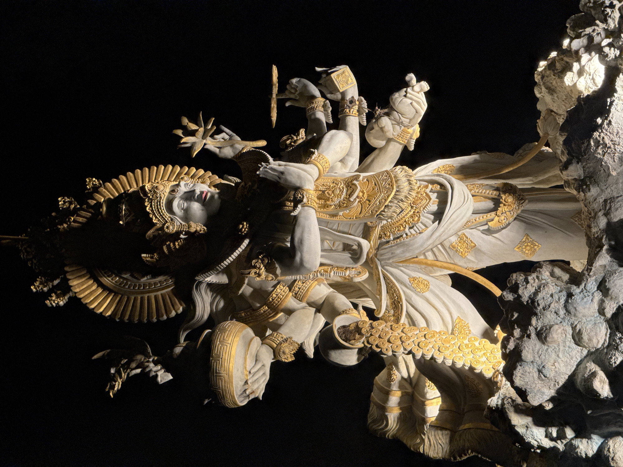 The image depicts a statue of a reclining figure with multiple arms, adorned with gold accents. It is located in North Kuta, Indonesia, adjacent to a rock formation. The figure appears to be lying down with its head to the left. Its face is pale with red lips. It wears a large headpiece with golden rays emanating outwards. The arms are arranged in various positions, holding objects like a lotus flower and a disk. The body is draped in ornate, white and gold garments. The background is black, with the exception of the rock formation on the right side of the image.