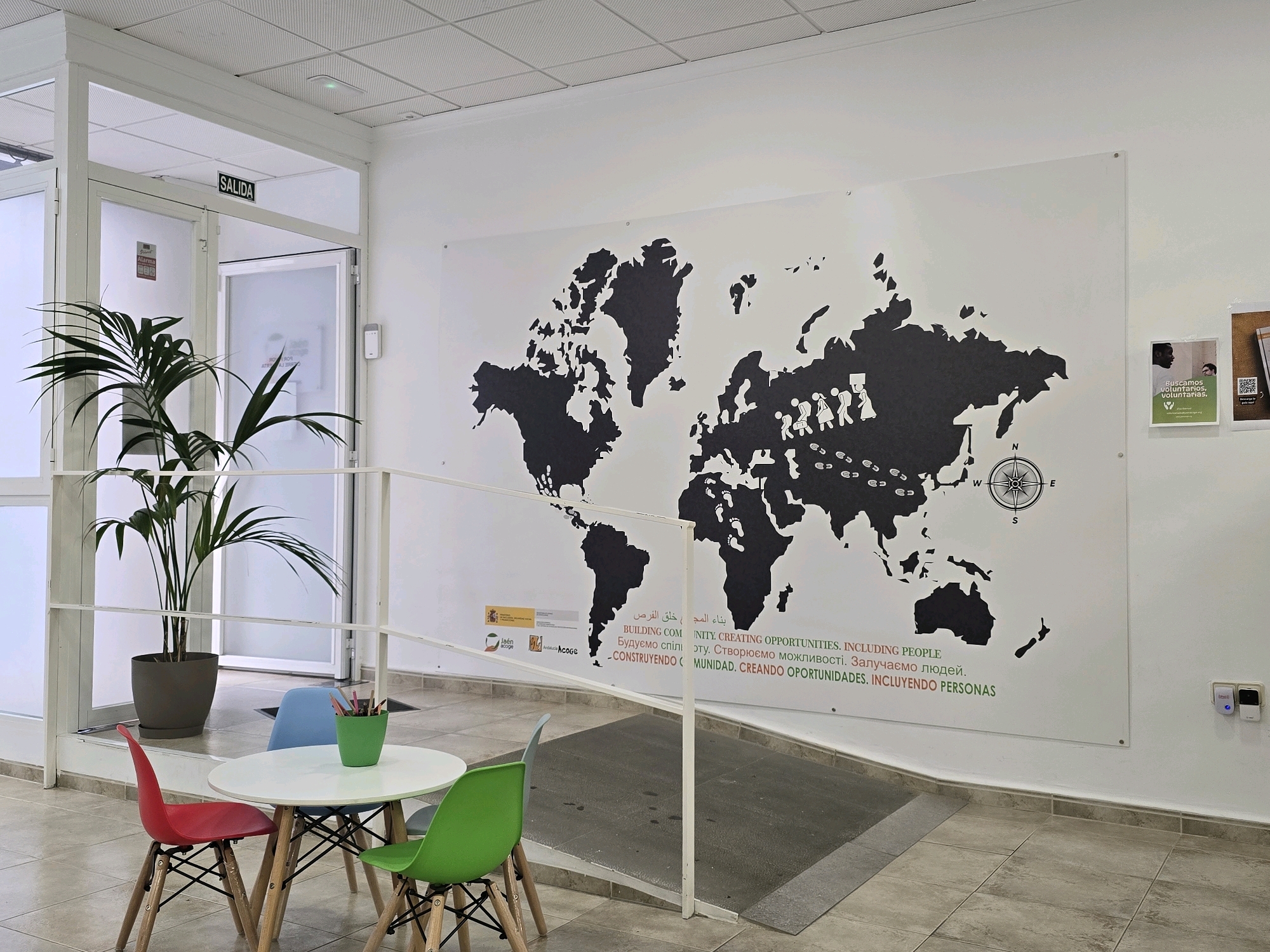 The image depicts an interior scene, likely an office or community space, situated in Jaén, Spain. Prominently displayed on the white wall is a large black world map mural. The map features stylized figures of people walking across continents and footprints. Below the map, there is text in multiple languages, including "BUILDING COMMUNITY. CREATING OPPORTUNITIES. INCLUDING PEOPLE." and "CONSTRUYENDO COMUNIDAD. CREANDO OPORTUNIDADES. INCLUYENDO PERSONAS." To the left of the mural, a large potted plant with long, green leaves stands next to a glass door with a "SALIDA" (EXIT) sign above it. In the foreground, a small, round white table is surrounded by three child-sized chairs: one red, one blue, and one green. A green pot filled with colored pencils sits on the table. The floor is tiled in a neutral, earthy tone. In the background, other doors and office elements are visible, along with promotional posters on the right wall. The overall impression is one of a welcoming and globally-minded environment.
