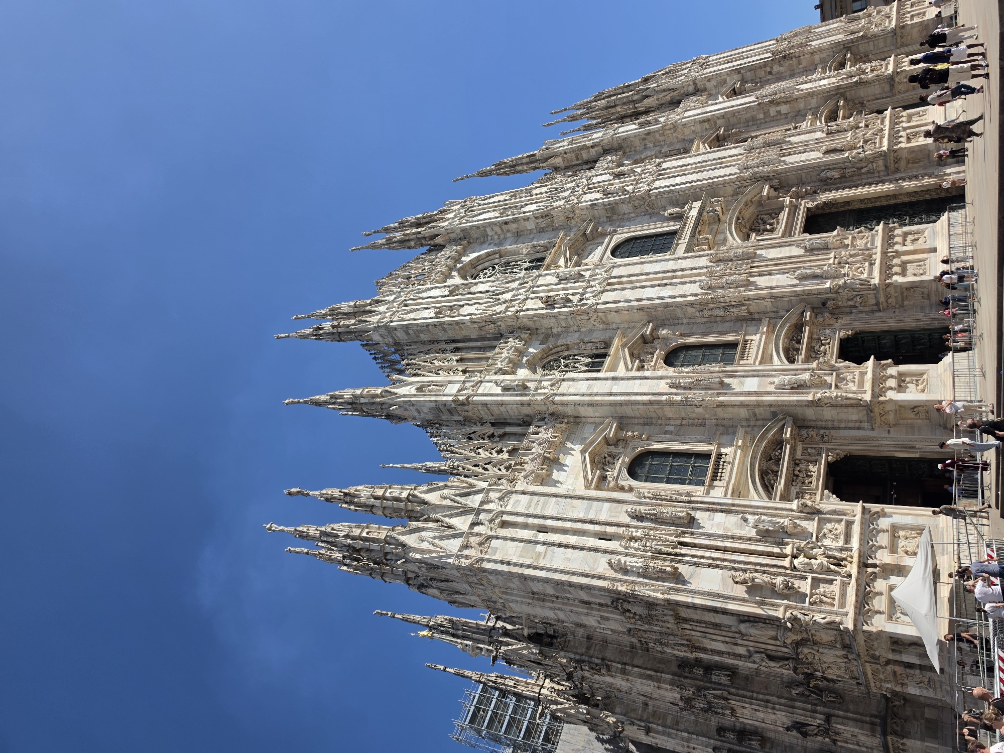 The image shows the Milan Cathedral, also known as the Duomo di Milano, a Gothic cathedral located in Milan, Italy. The cathedral is characterized by its intricate facade, adorned with numerous spires, statues, and architectural details. The building is constructed primarily of white marble, which reflects the sunlight, giving it a bright and majestic appearance. The sky above is a clear, deep blue, with a few faint clouds visible.
In the foreground, people are gathered, some standing near the entrance to the cathedral, while others are walking on the plaza. There are barriers set up, possibly for crowd control or to guide visitors. A white tent is set up to the left of the main entrance. On the left side of the image, scaffolding is visible on a portion of the cathedral, suggesting ongoing maintenance or restoration work. The overall scene depicts a vibrant public space in front of a renowned historical landmark on a sunny day.