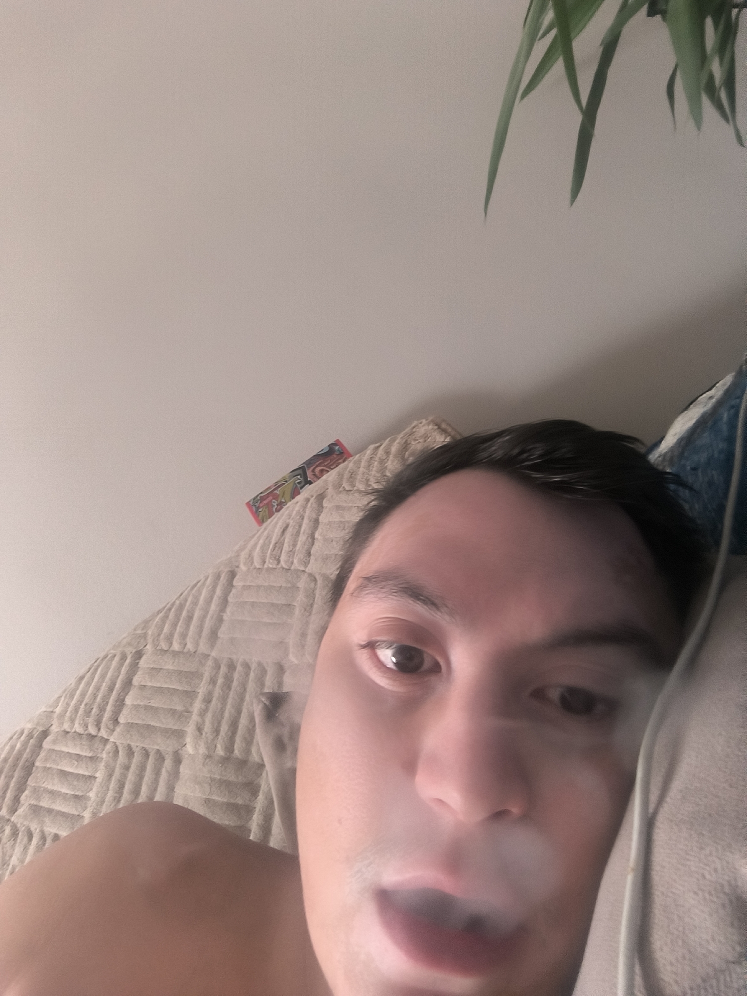A close-up, low-angle shot shows a person lying on a textured beige blanket, with a pillow to the right. The person's face is partially obscured by a cloud of smoke. The background is a plain, light-colored wall, with a hint of a plant's leaves visible in the upper right corner. A colorful, rectangular object, possibly a small box or wrapper, is positioned on the blanket near the person's head. The scene is indoors. There is no indication of the city of Kovrov, Russia.