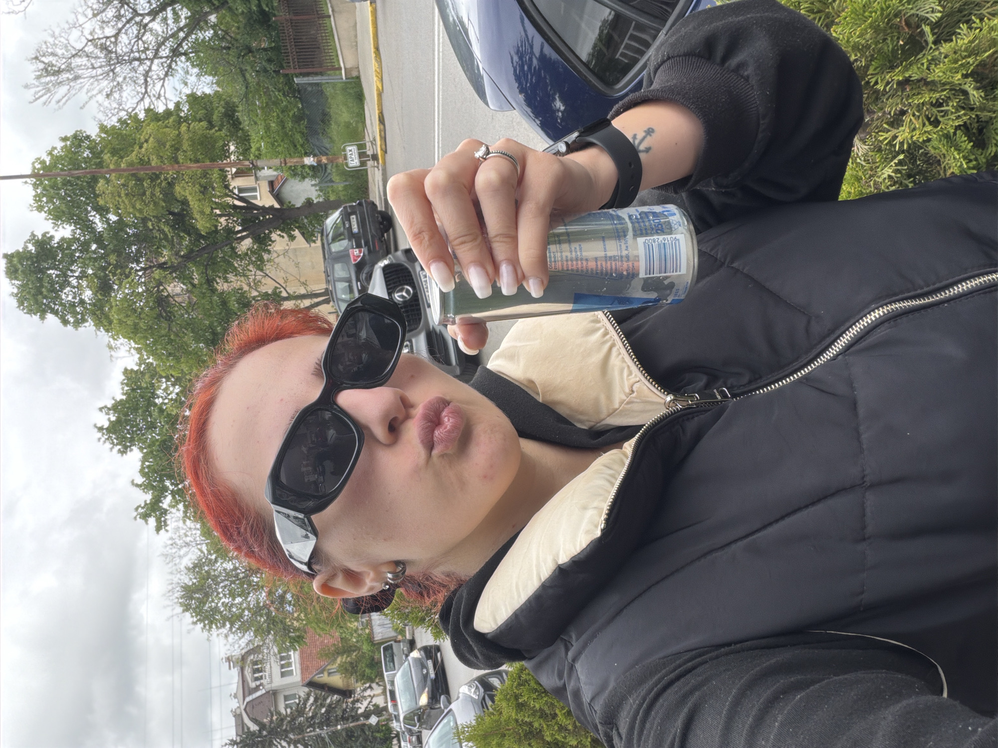 A person with red hair and sunglasses is holding a can of Red Bull. The person is wearing a black puffer vest over a black hooded sweatshirt, and has a ring and watch on their left hand. The background includes trees, buildings, parked cars, and street signs. The location is likely Sofia, Bulgaria, as indicated by the Cyrillic script on the street sign.
