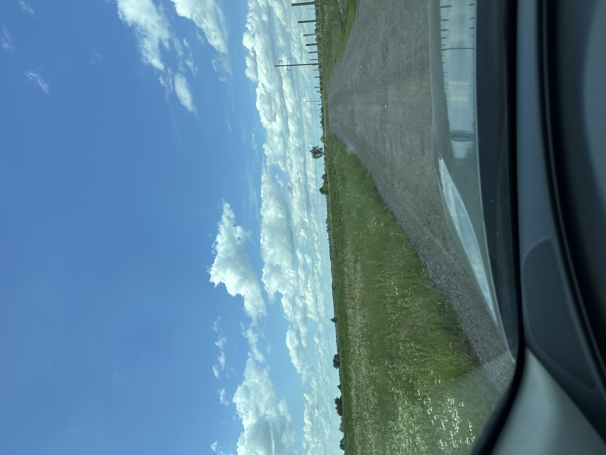 The image shows a view from inside a vehicle looking out at a dirt road stretching into the distance. To the left of the road is a field of green grass with some white wildflowers. To the right of the road, a fence runs along the edge. In the background, there are trees and utility poles under a bright blue sky with fluffy white clouds. The scene appears to be in a rural area.