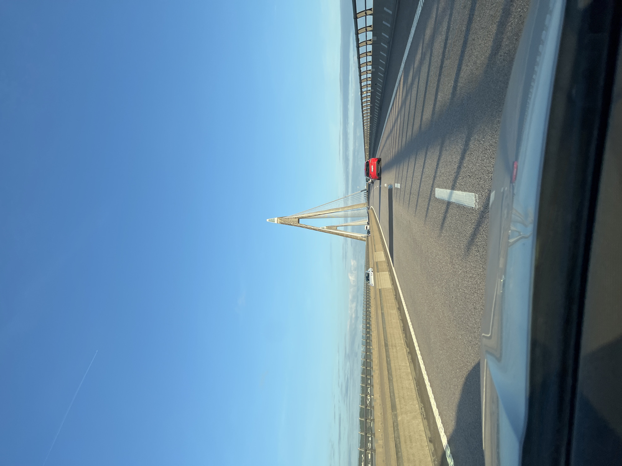 The image depicts a view from a vehicle driving on a highway across a large bridge. The bridge has multiple lanes of traffic separated by white lines. On the right side of the road, there is a metal railing. In the distance, a prominent cable-stayed bridge tower is visible, with cables extending from it to support the bridge deck. A red car is in the foreground, traveling in the same direction as the camera. The sky is clear and blue with some thin clouds near the horizon. The scene appears to be well-lit, suggesting it is daytime. There is no information in the image to suggest it is in Herrestad, Sweden.