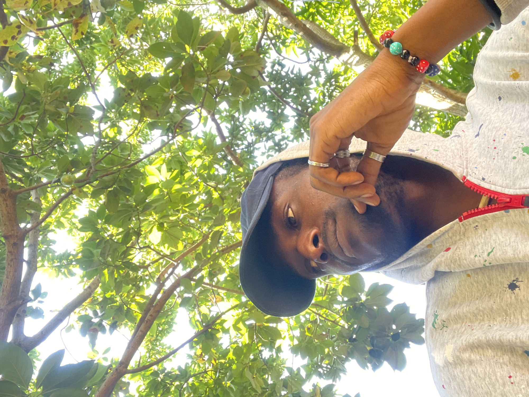 In Jos, Nigeria, a low-angle shot captures a young Black man outdoors, beneath the dappled shade of lush green trees. He wears a black baseball cap and a grey hooded sweatshirt adorned with colorful paint splatters. His right hand is raised to his face, fingers lightly touching his cheek, and he wears two silver rings. A bracelet on his wrist features a pattern of red, blue, and turquoise beads, some resembling dice. The man's gaze is directed upwards and slightly to his right, suggesting he is looking at something above or beyond the frame. The sunlight filters through the leaves, creating a bright, airy atmosphere.
