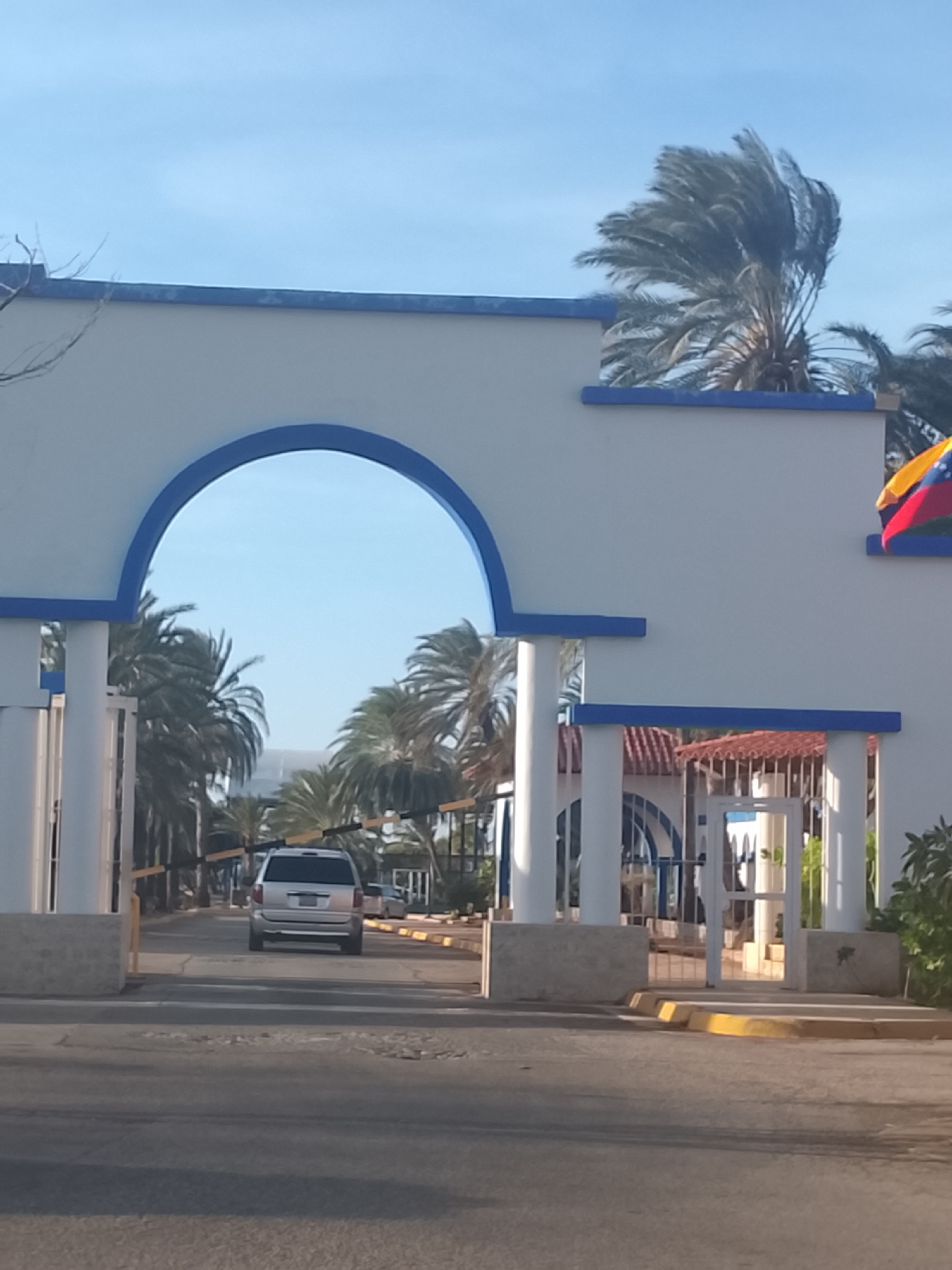 The image shows a white and blue archway entrance leading to a road lined with palm trees, with a silver minivan driving through the arch. A Venezuelan flag is attached to the archway structure on the right. Buildings with red roofs are visible behind the arch. The location is likely Altagracia, Venezuela based on the architectural style, vegetation, and national flag.