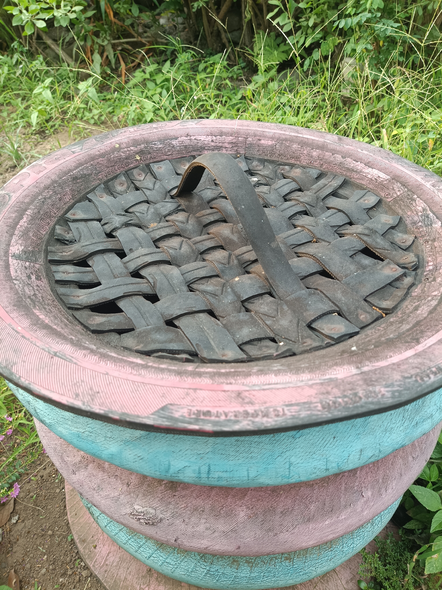 The image shows a close-up view of a planter made from stacked tires. The top tire is painted pink and has a woven cover made from strips of black rubber, possibly from another tire, with a black handle attached. The underlying tires are painted in alternating bands of pink and teal. The planter is situated outdoors, surrounded by green foliage and soil. The scene is located in the city of Jember, Indonesia. There are no people visible in the image.