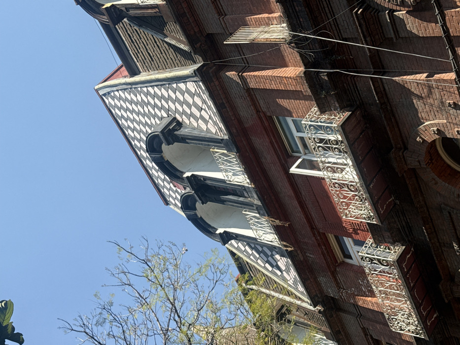 The angled view shows a building with intricate architectural details. It features black and white diamond-patterned facades, arched balconies with white railings, and brick walls. Windows are framed with ornate white ironwork. Parts of a tree are visible at the building's base, set against a clear blue sky. The overall style appears historic and detailed.