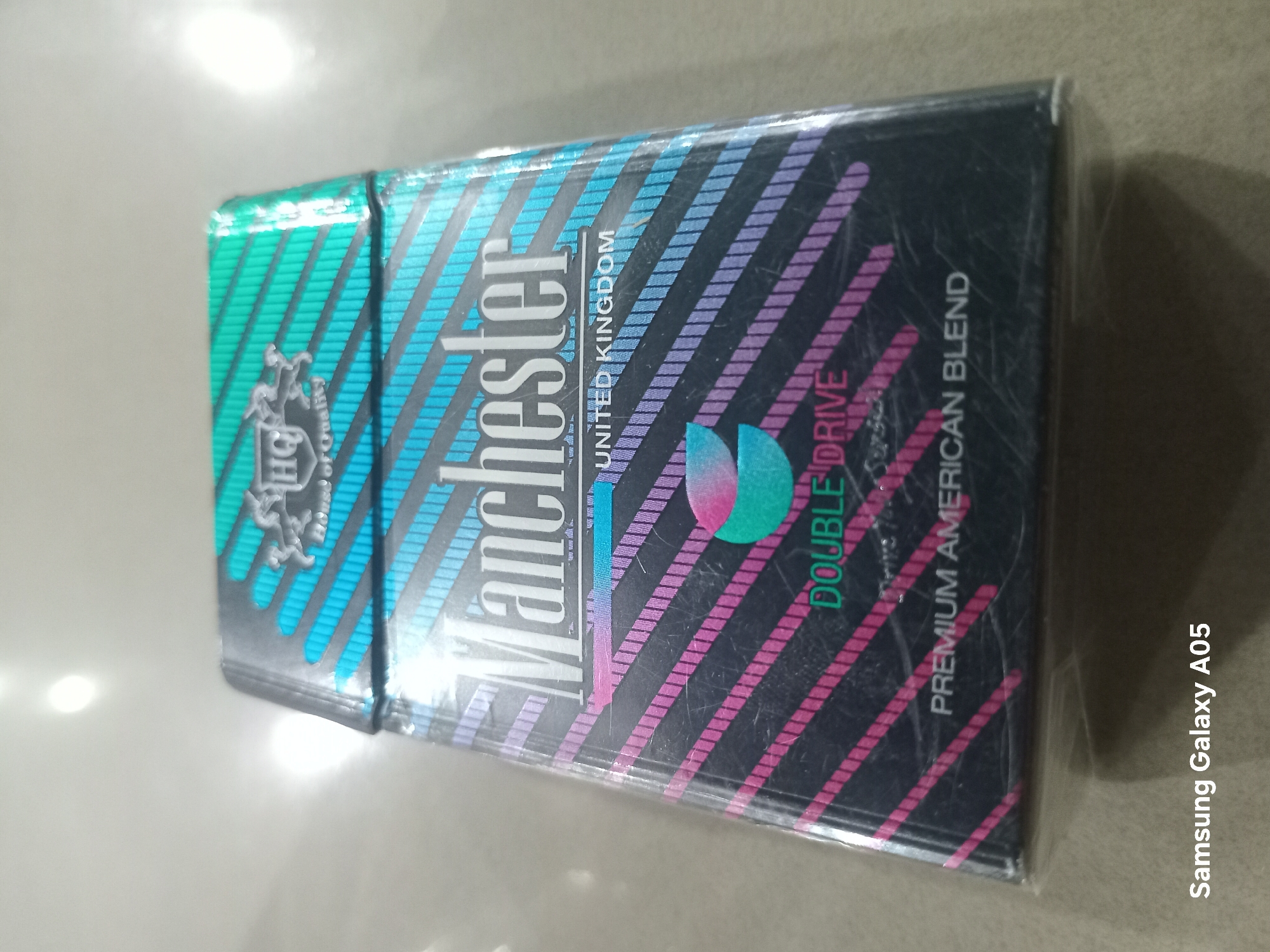 The image captures a single pack of Manchester brand cigarettes, still sealed in its clear plastic wrapping, lying on a light-colored, reflective surface. The cigarette pack features a striking design with a black background accented by diagonal holographic stripes. These stripes transition from shades of blue and teal on the left to pink and purple on the right.
The brand name "Manchester" is prominently displayed in large white letters across the center of the pack. Below this, positioned vertically, is the text "UNITED KINGDOM," indicating the product's country of origin. Further to the right, a small circular emblem, blending teal and pink colors, accompanies the words "DOUBLE DRIVE," suggesting a specific flavor or variant. Below this, "PREMIUM AMERICAN BLEND" describes the tobacco type. On the left side of the pack, a silver crest or emblem with the letters "HQ" and the phrase "of Quality" is visible.
The surface on which the pack rests is smooth and reflects a bright light source, causing a glare in the upper left portion of the image. The plastic wrapper shows minor creases and reflections. The scene is static, depicting only the product. The image contains a "Samsung Galaxy A05" watermark in the bottom right corner. While the location context is Lhokseumawe, Indonesia, there are no specific visual cues within the image itself to confirm this geographical detail.