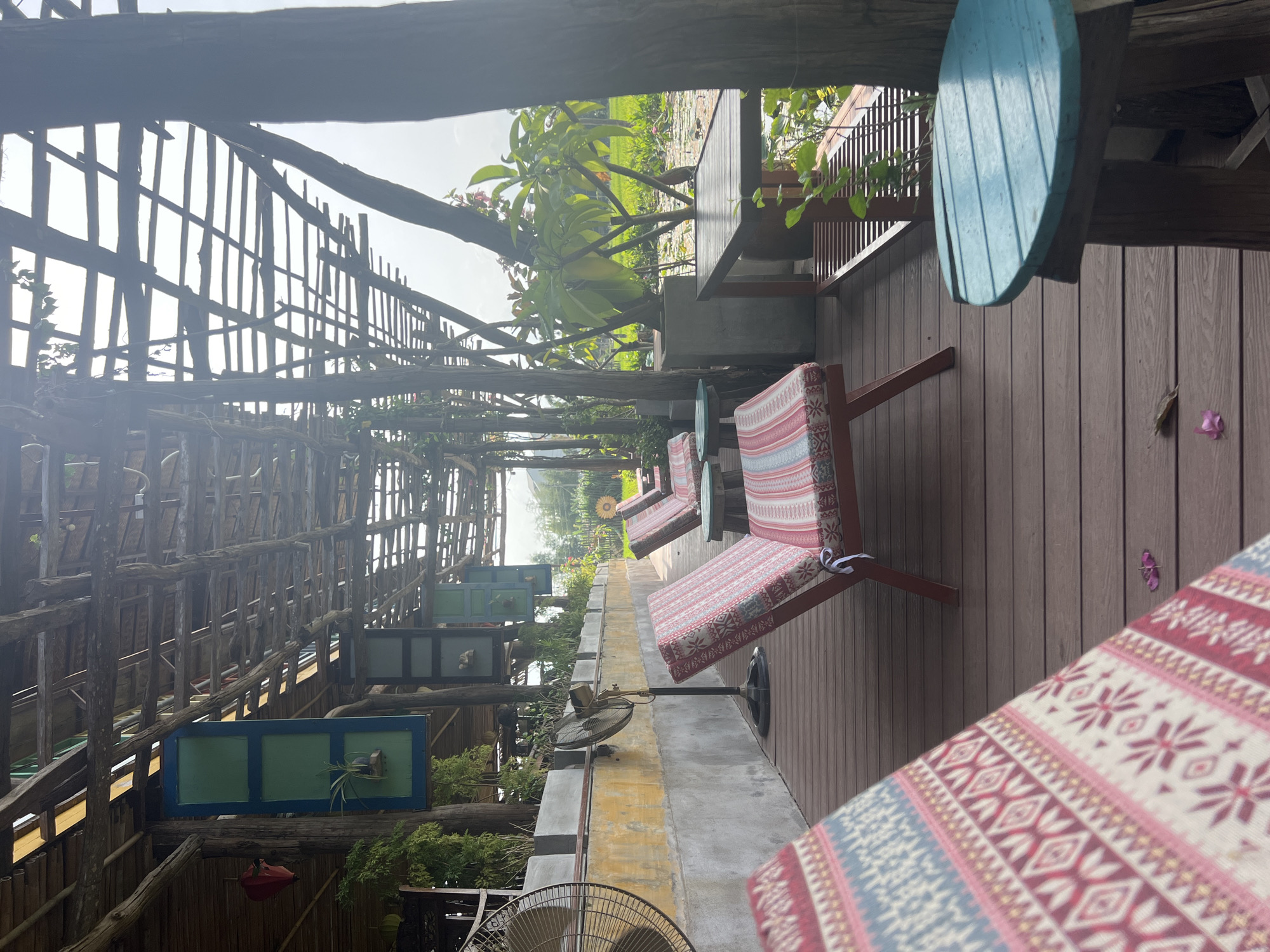 An outdoor seating area overlooks a lush green landscape, possibly a rice paddy or a field with water. The seating area is made of wooden planks and features several chairs with colorful patterned cushions. Some of these chairs are in the foreground, while others recede into the background. In the middle ground, there are two standing fans, one larger and black on the left, and a smaller, gold-colored one on the right. Behind the chairs, a low concrete wall with a yellow-painted section runs along the back. Above this wall, plants are arranged in planters. The entire area is covered by a rustic wooden roof structure supported by wooden posts. Decorative elements, such as hanging lanterns and repurposed window frames with plants, are visible attached to the overhead structure. A wooden table with a slatted shelf is positioned to the right, with a small, round, turquoise-colored table in front of it. The natural light suggests it is daytime. This scene is characteristic of a tranquil, nature-oriented establishment in Hoi An, Vietnam, likely a cafe or a guesthouse situated near a rural setting.