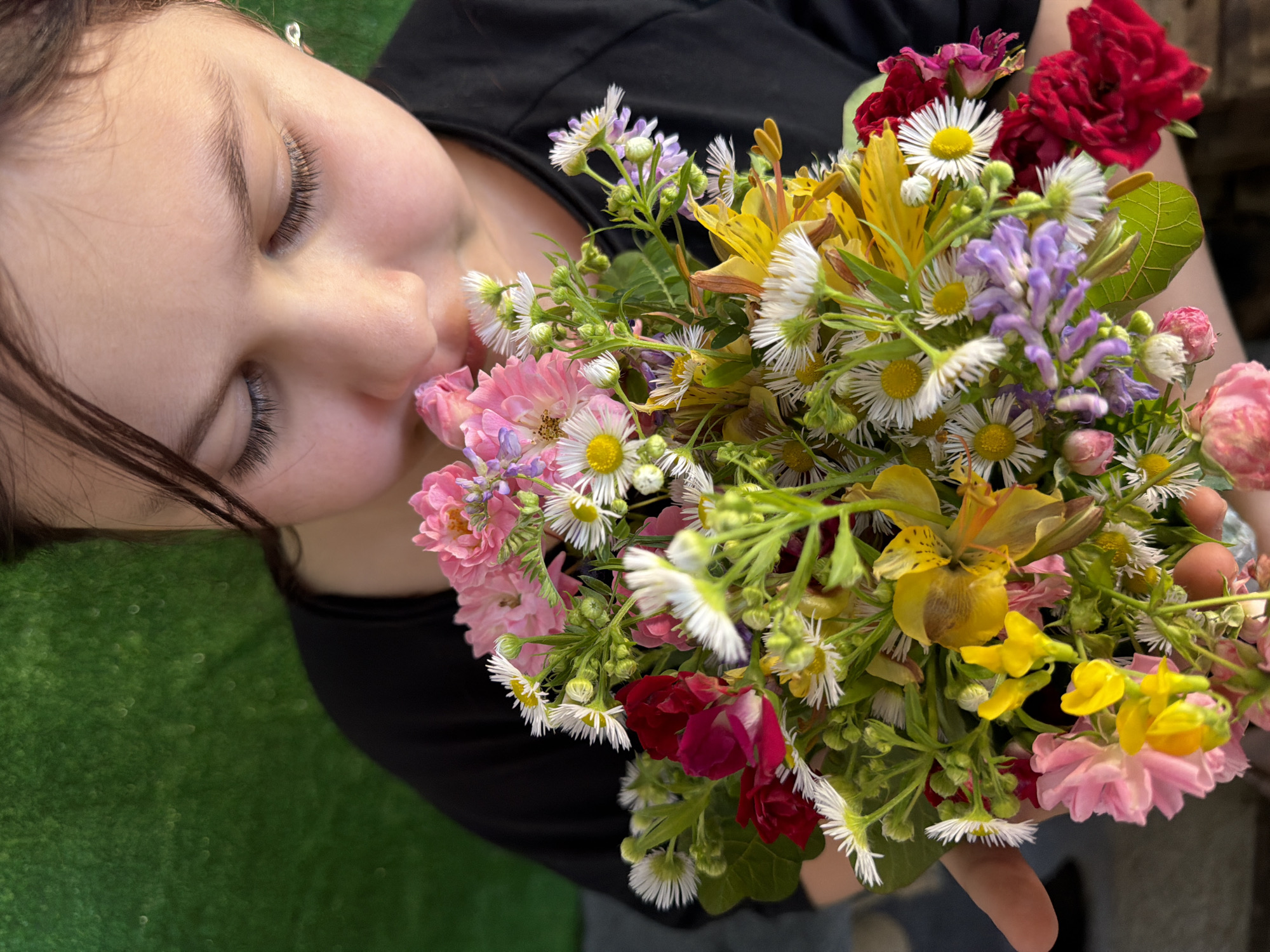 A young person in a black t-shirt is holding a bouquet of wildflowers and smelling them. The flowers are a mix of colors, including pink, white, yellow, and red. The background is a blurred green texture, possibly a wall or fabric. The lighting is soft and natural. There is no visible indication of the city of Tuapse, Russia in this image.