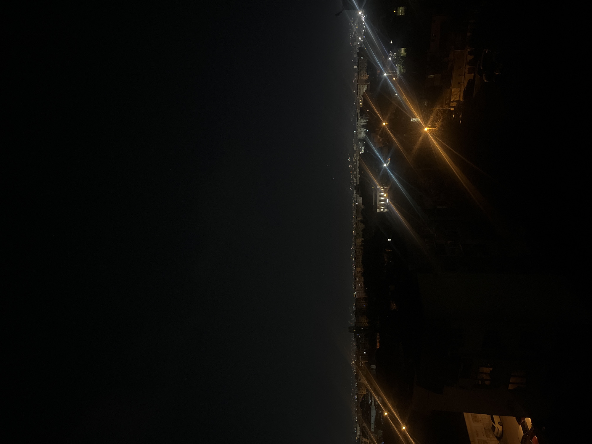 The image shows a night cityscape. The right side of the image depicts a street with lit streetlights and buildings. The left side of the image is a dark, seemingly open space, possibly water or a dark field. The overall scene is dark with points of light from the city. It is not possible to determine if this is Sofia, Bulgaria or any other specific place.