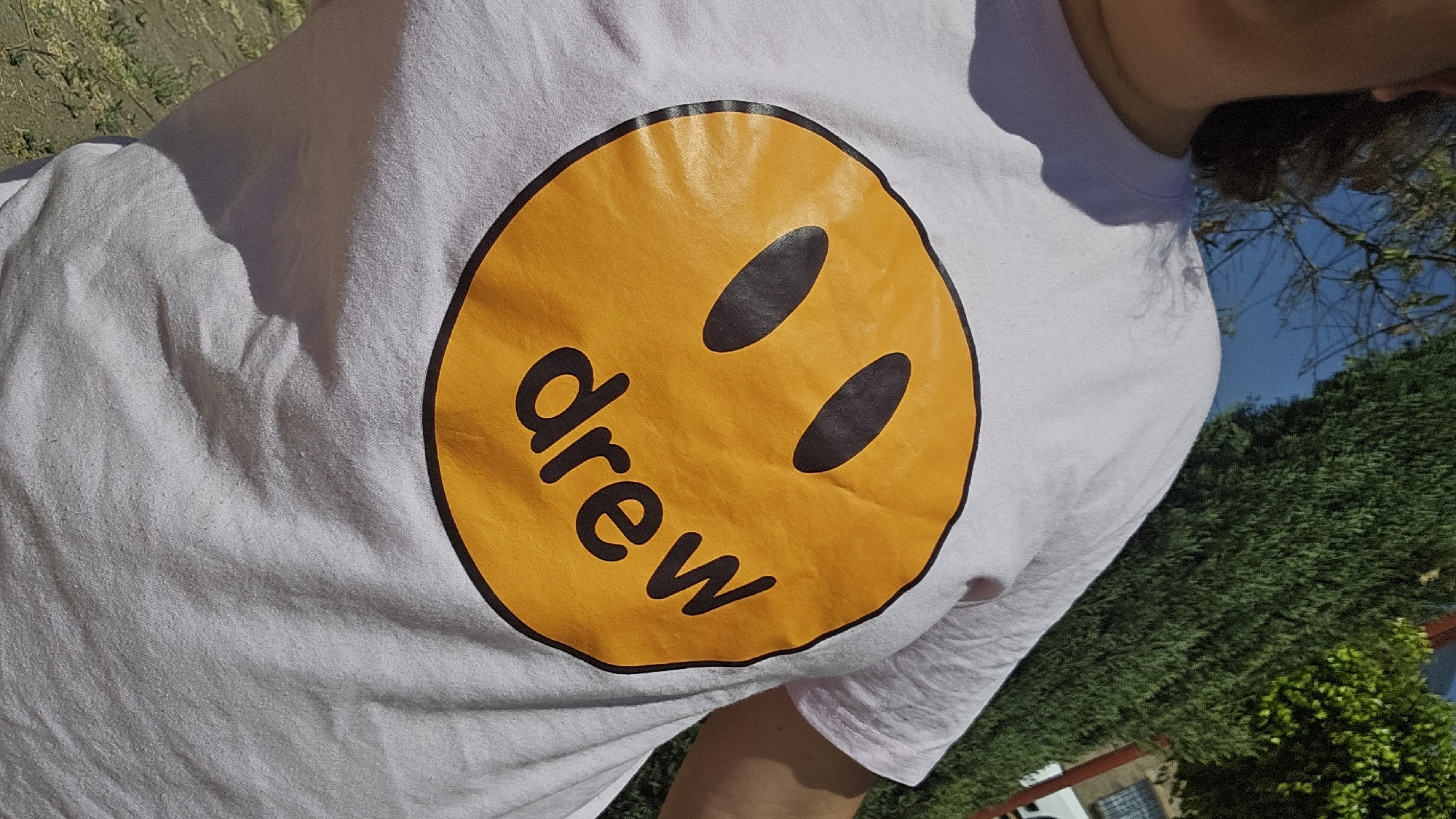 The image captures a close-up, waist-up view of an individual outdoors in San Bernardino Tlaxcalancingo, Mexico. The person's lower face, neck, and left shoulder are partially visible. They are wearing a light lavender t-shirt featuring a prominent, large circular yellow graphic on the chest. This graphic depicts a smiley face with two black oval eyes and the lowercase word "drew" in black, positioned as the mouth. The t-shirt fabric shows evidence of pilling. The outdoor environment includes a clear blue sky visible in the upper left, with dark, bare tree branches silhouetted against it. To the right, a tall, dense green conifer hedge extends vertically. Beyond the hedge, a building with a visible window and partial structural elements can be discerned. The ground in the lower background, beneath the person's right side, consists of dry earth with sparse green vegetation.