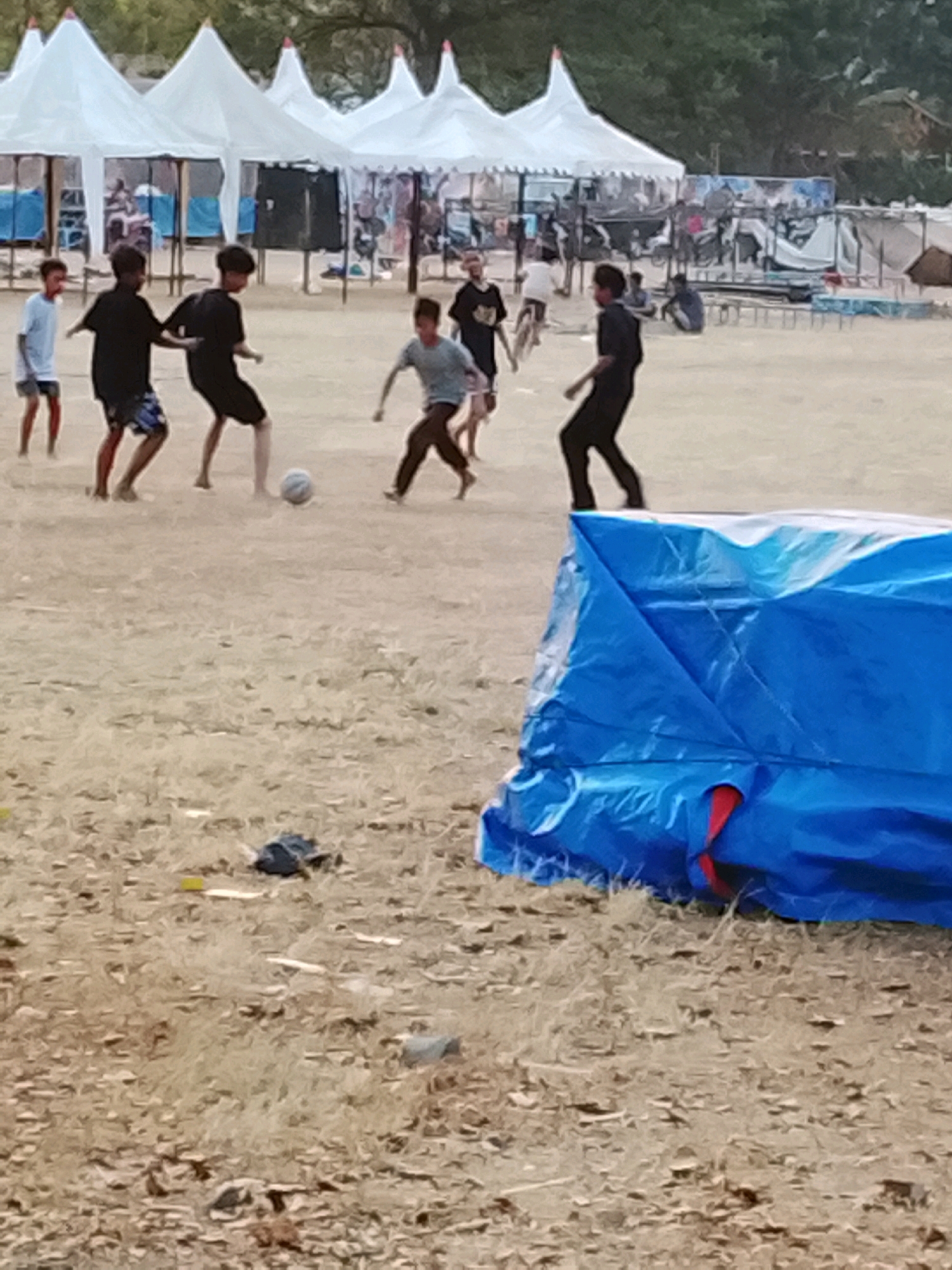 In Harjosari Lor, Indonesia, a group of young boys are playing soccer on a dusty field. Several white tents are pitched in the background, suggesting a temporary settlement or event. The overall scene depicts a casual outdoor activity amidst a somewhat sparse environment.