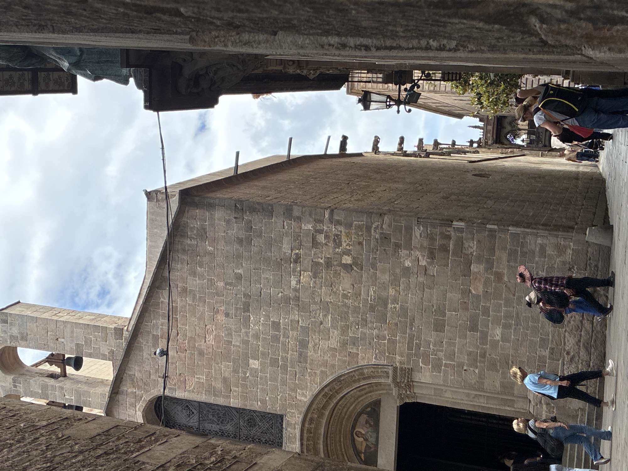 This eye-level photo depicts a scene in Barcelona, Spain, specifically the entrance to the Basilica de Santa Maria del Pi. The perspective is from within a narrow stone passageway, framing the basilica's facade.
The facade is constructed of large, tan stone blocks and includes an arched doorway, a tall bell tower, and a rose window covered with a metal grate. Several people are walking in front of the basilica, including tourists with backpacks.
The sky is visible in the background, with light white clouds. The foreground is in shadow due to the narrow passageway. Elements of the surrounding buildings are visible at the top of the frame, partially obscuring the view of the sky.