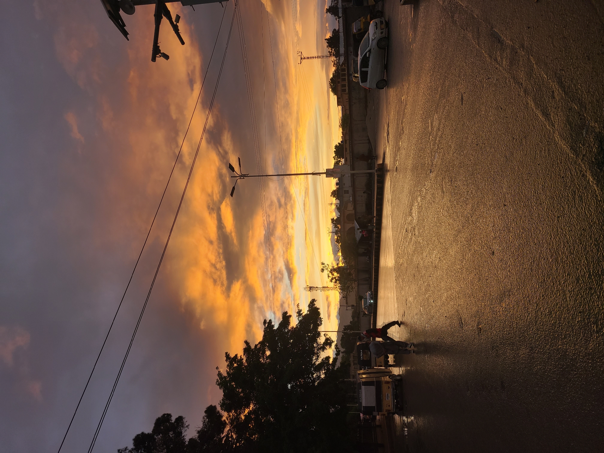 The image depicts a street scene at sunset in Quetta City Tehsil, Pakistan. The sky is filled with orange and yellow clouds, contrasting with darker purple and gray clouds near the horizon. Electrical lines are visible against the sky. On the right side of the image, there's a wet street reflecting the golden light. A parked white car is visible on the side of the road. In the foreground, figures of people and vehicles, likely auto-rickshaws, are silhouetted against the bright street. Trees line the road. Streetlights are also visible.